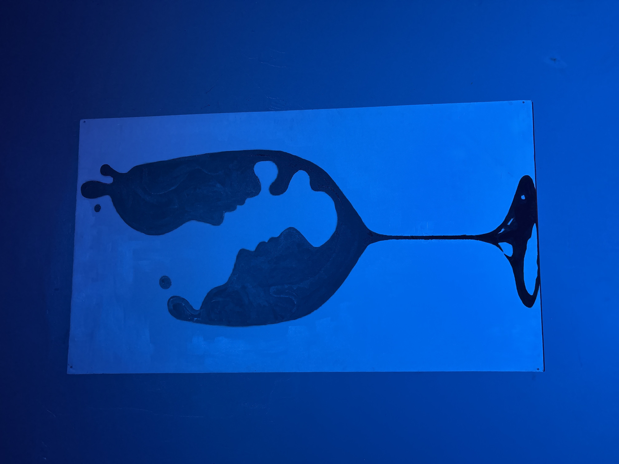 An abstract art piece is displayed on a wall, featuring a predominantly blue palette with a dark, amorphous shape resembling a spilled drink or a Rorschach inkblot. The shape vaguely resembles a wine glass, with a narrow stem and a wider, irregularly formed base. Within the shape, it is possible to discern the profiles of two faces seemingly positioned face-to-face. The artwork is mounted on a rectangular backing board, which is secured to the wall with screws or similar fasteners visible at the corners. The surrounding wall is painted a solid, matching shade of blue, creating a monochromatic backdrop for the artwork. The artwork is located in the city of Lafia, Nigeria.