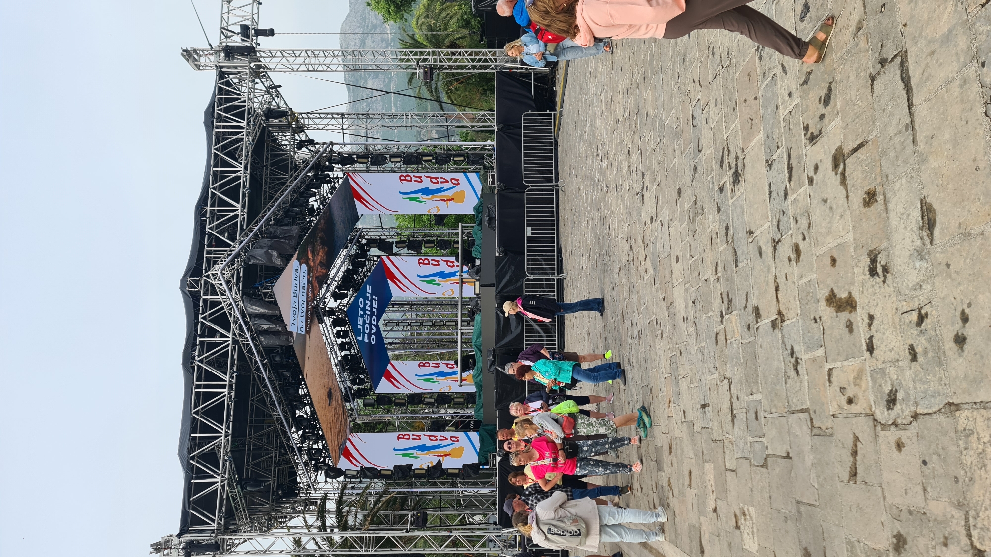 The image shows an outdoor scene with a large stage structure dominating the view. The stage is constructed of metal scaffolding, covered in black draping, and adorned with promotional banners with the word "Budva" prominently displayed, indicating a location in Budva, Montenegro. Several people are standing in front of the stage, appearing to be a group or audience. The environment includes stone pavement and some greenery in the background, suggesting a public square or event space. Some of the stage’s lighting fixtures are visible. The atmosphere appears to be during daylight. A section of metal barriers is also seen near the stage. Some palm trees are also present in the image.