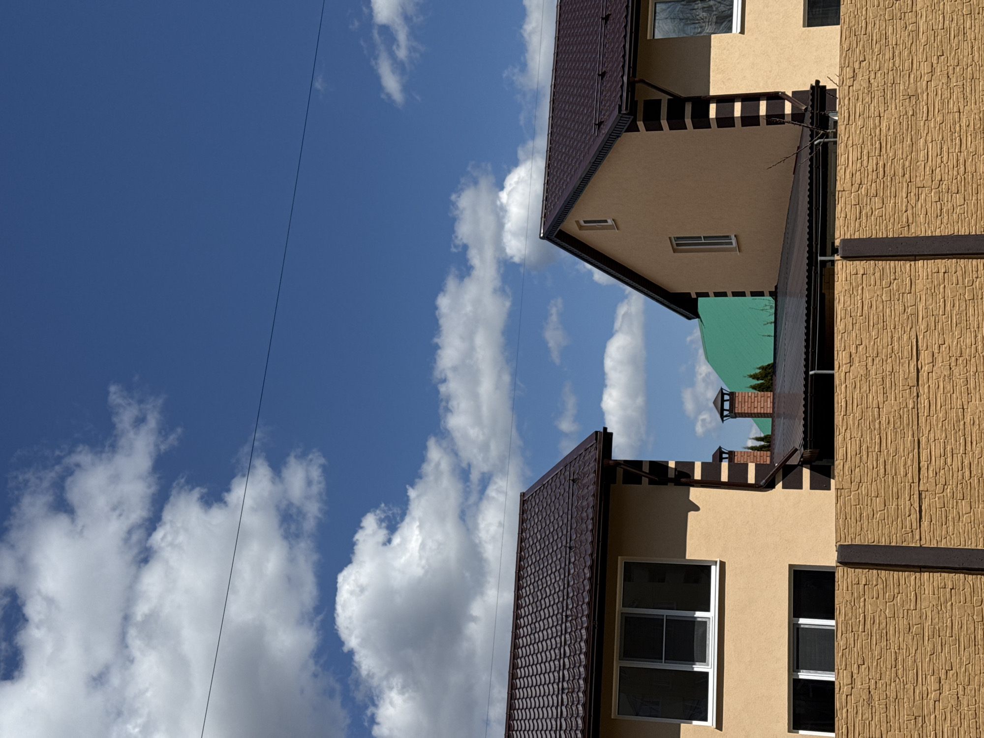 The image captures an outdoor scene featuring sections of multiple buildings against a bright blue sky with scattered white clouds. The perspective is tilted, with the sky occupying the upper and left portions of the frame, and buildings along the bottom and right.
Two prominent buildings with tan or light brown facades are visible. The building on the left has a dark brown tiled roof and tan walls with several windows, some featuring white frames. Below the roofline, there is a decorative section with vertical dark brown and tan stripes. The building on the right presents a textured, rough-hewn tan facade, accented by a dark brown horizontal band, with a few windows also visible.
In the background, nestled between these two structures, another building is partially visible, distinguished by a bright green roof and a brick chimney with a dark brown cap. A thin, dark utility wire stretches diagonally across the sky. The scene is brightly lit by the sun, suggesting it is daytime, likely morning or afternoon, as evidenced by the clear sky and visible shadows. There are no people or specific activities depicted. No text is visible within the image.
