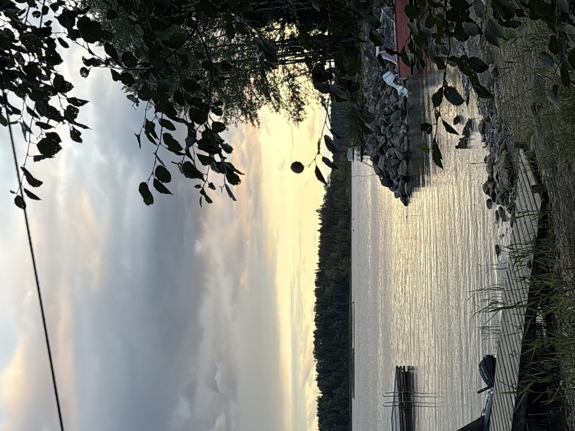 This is a photograph of a lake in Rautalakhti, Russia, at sunset. The sky is filled with clouds, with hints of yellow and pink from the setting sun. The lake is calm, with ripples reflecting the sky. In the foreground, there is a wooden dock with a small boat and an outboard motor. To the right, there is a rocky shoreline with trees and bushes. In the distance, across the lake, a dense forest borders the water. There are no people visible in the image.