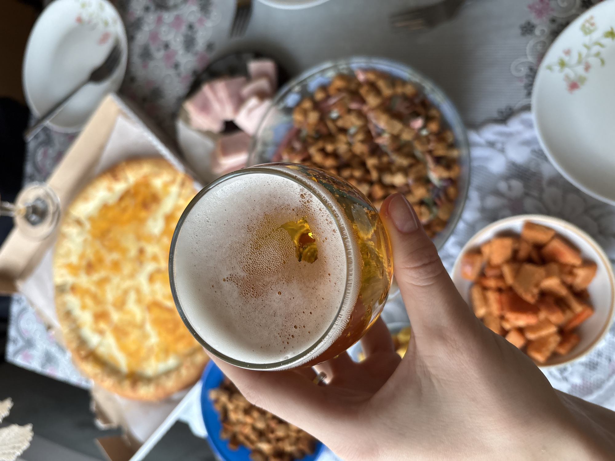 A hand holds a glass of beer above a table laden with food. The table is covered with a patterned tablecloth and has several plates and bowls containing various dishes. There's a round cheese-filled flatbread in a cardboard box, a plate with sliced ham, a bowl of fried croutons, and another bowl with cubed, orange food. A white plate with a floral design and a fork are also visible. The setting seems to be a casual meal or gathering. The location, Ust-Ilimsk, Russia, is provided context, but not visible within the image.