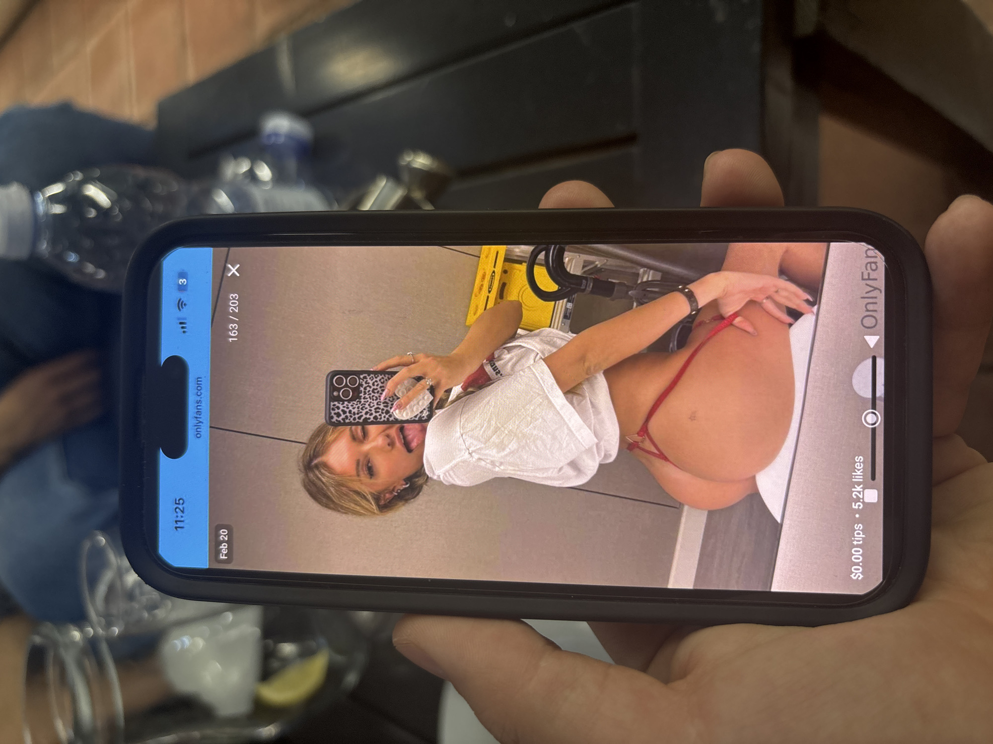 A right hand holds a black smartphone, displaying an image from "onlyfans.com" dated "Feb 20" at "11:25". The screen shows a young woman with blonde hair taking a mirror selfie. She winks and sticks out her tongue, holding a leopard-print smartphone with her right hand. She wears a white cropped t-shirt and a red thong, with her left hand positioned on her left buttock, pulling the thong downwards. The background of the displayed image features light-colored walls, a yellow object (possibly equipment), and black hoses. The bottom of the phone screen shows "$0.00 tips • 5.2k likes" and partial text "OnlyFan". Behind the held smartphone, out of focus, are elements of a real-world setting: a dark table, a clear glass pitcher with a lemon wedge and ice, an empty glass, a plastic water bottle, and reddish-brown tiled or paved flooring. Two blurry figures are seated in the far background. The scene is located in the city of Blagoevgrad, Bulgaria.