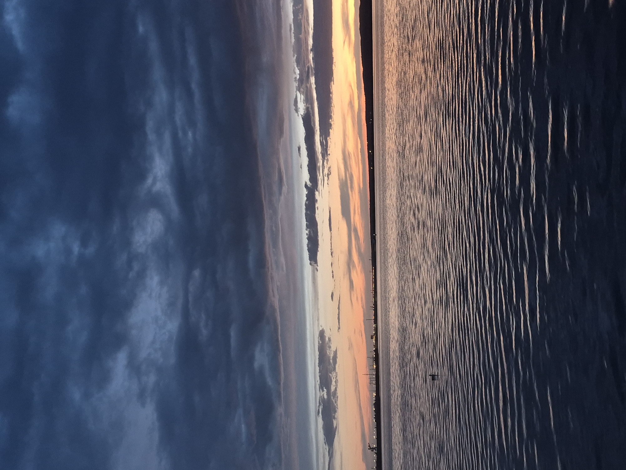 The image depicts a sunset over water, likely the Kama River in Chaikovsky, Russia. The sky is split into three main sections: dark blue clouds on the left, a transition of white and orange clouds in the middle, and a bright orange sky on the right where the sun has set. Below the horizon line, a body of water is visible with small waves reflecting the colors of the sky. Several small lights can be seen in the background on the shoreline.