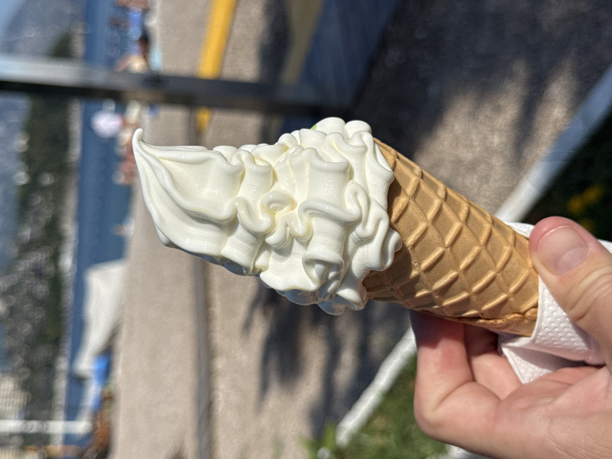 This is a close-up, handheld photograph of a person holding a vanilla soft-serve ice cream cone. The ice cream is swirled high on the cone, with a slightly green tip visible at the very top. The cone itself is a classic waffle cone with a diamond pattern. The person's hand, covered by a white paper napkin, is visible at the bottom right of the frame, holding the cone. The background is blurred, suggesting an outdoor setting on a sunny day. In the distance, a body of water, possibly the sea, is visible along with some distant buildings and greenery. There are also hints of people in the background, adding to the sense of an active, public place. The overall impression is one of a casual, summery moment. The lighting suggests it is daytime. The location context of Budva, Montenegro, implies a coastal setting, likely a tourist area given the presence of other people and the sea. There is no visible text in the image.