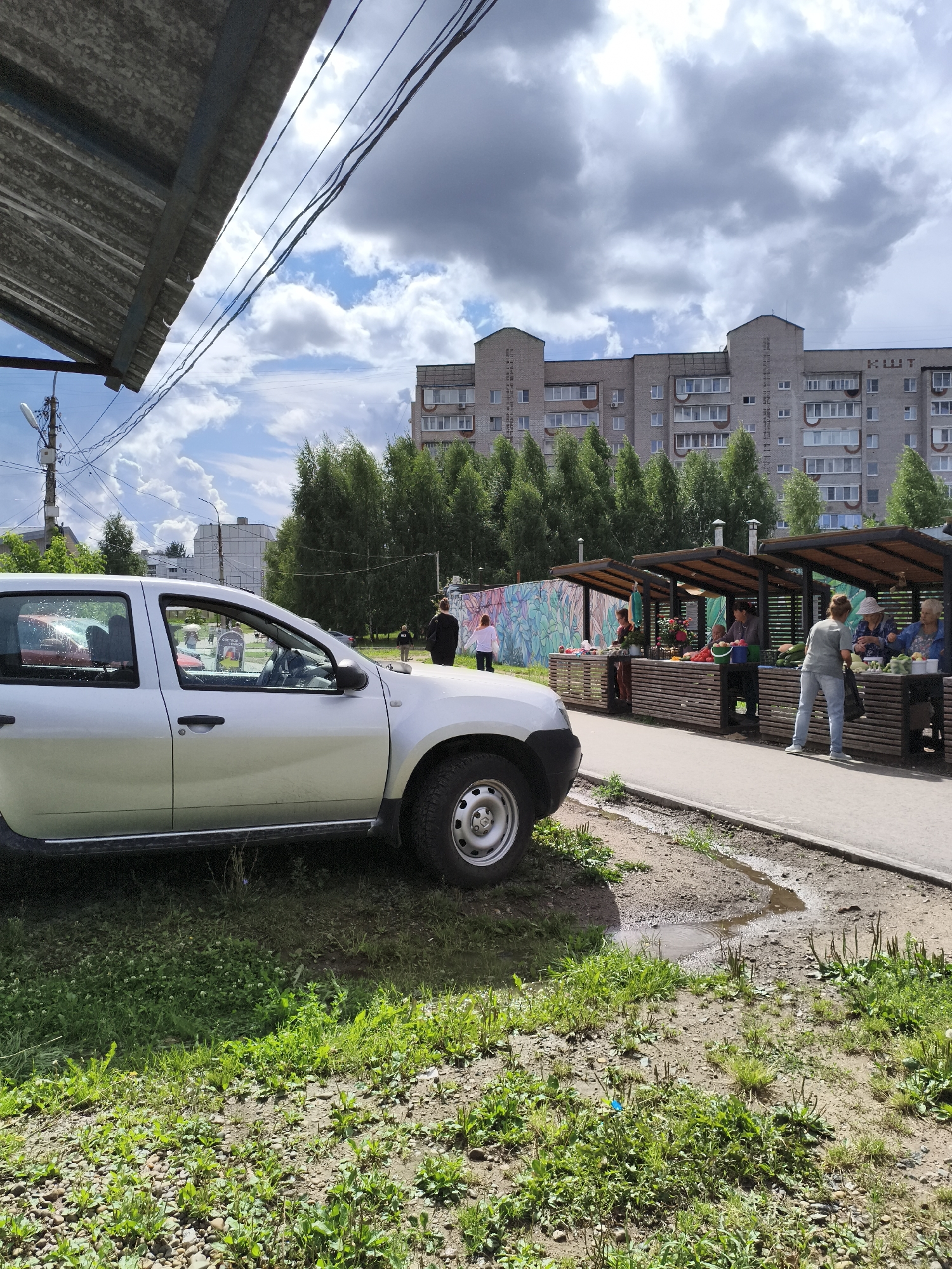 In the city of Chaikovsky, Russia, on a sunny day with scattered clouds, a silver car is parked on the left side of the frame. The car is facing right and takes up a significant portion of the foreground. Beyond the car, there is a market scene with several stalls set up, covered by awnings. People are visible at the stalls, appearing to be vendors and customers interacting. One stall has a sign that says "СЧАСТЛИВОЕ ВРЕМЯ" (Happy Time). The background features a multi-story brick apartment building and numerous trees. The ground in front of the stalls is paved, while the area where the car is parked and surrounding it is covered with grass and gravel, with some small puddles present. A mural depicting plant life is visible on a wall behind the stalls.