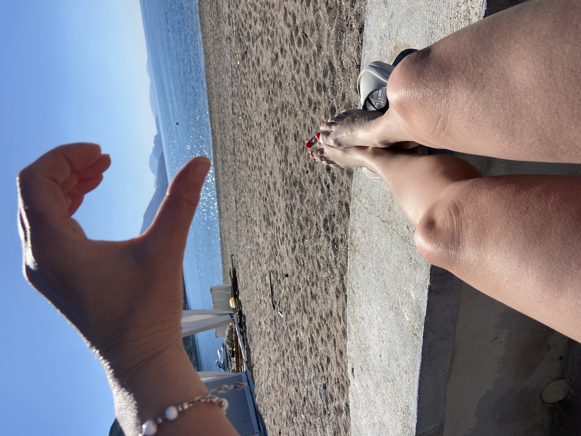 A beach scene in the city of Prijevor, Montenegro, is visible in an outdoor shot from a high angle. The subject is a person sitting on a concrete platform near a sandy beach, with their legs stretched out. The person is wearing sandals, and their toenails are painted red. The view extends to the blue sea and mountains in the background. A hand with a pearl bracelet on the wrist is raised, forming a small circle between the thumb and index finger. The sky is a clear blue, and sunlight is present, suggesting a sunny day.