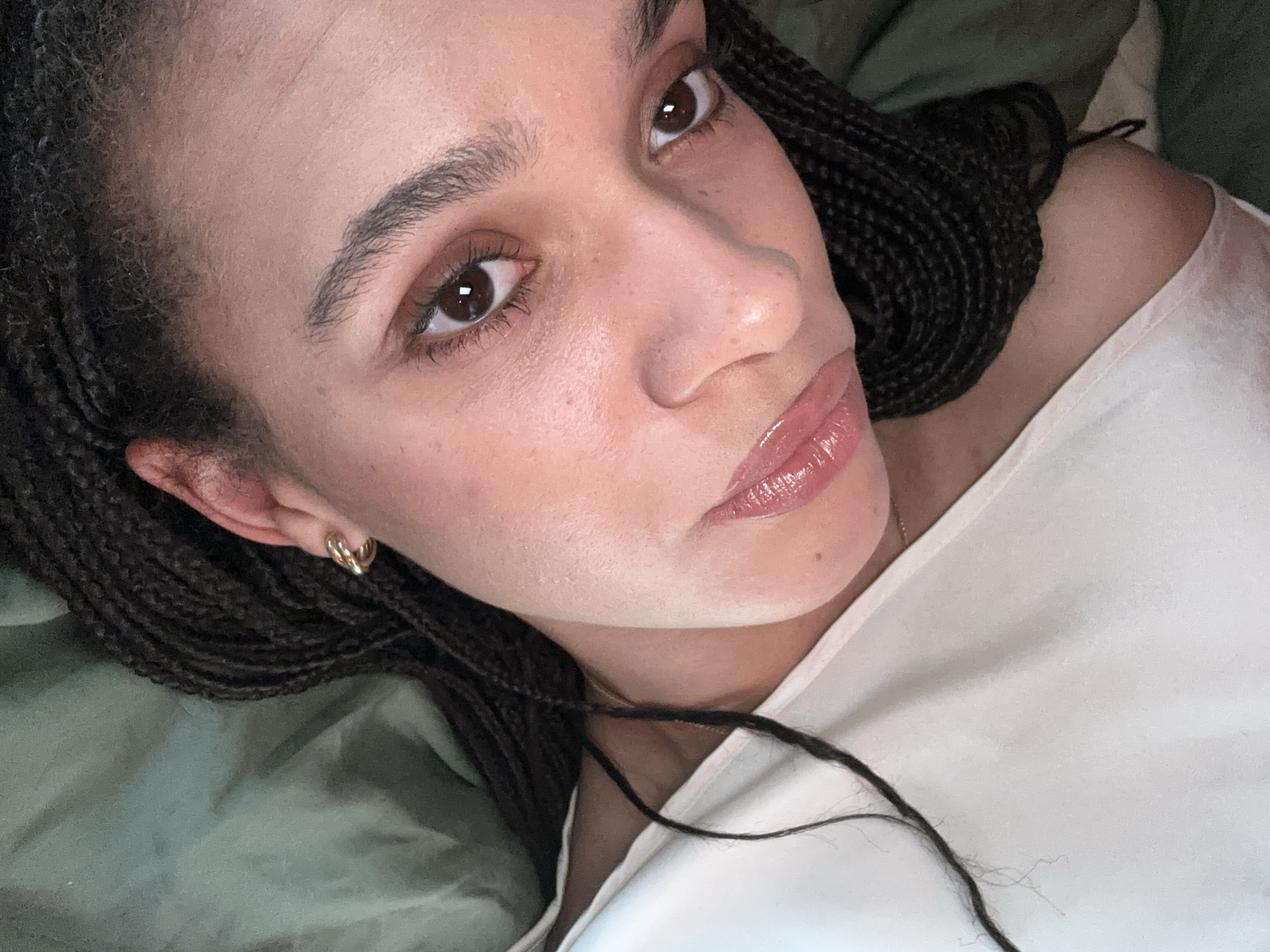 A close-up shot of a woman with braided dark hair, wearing a white top. Her skin appears smooth and clear. She has brown eyes, well-defined eyebrows, and glossy lips. She is wearing gold hoop earrings. The background is blurry and consists of green fabric. There is no indication of the location being in London, United Kingdom, or any other city.