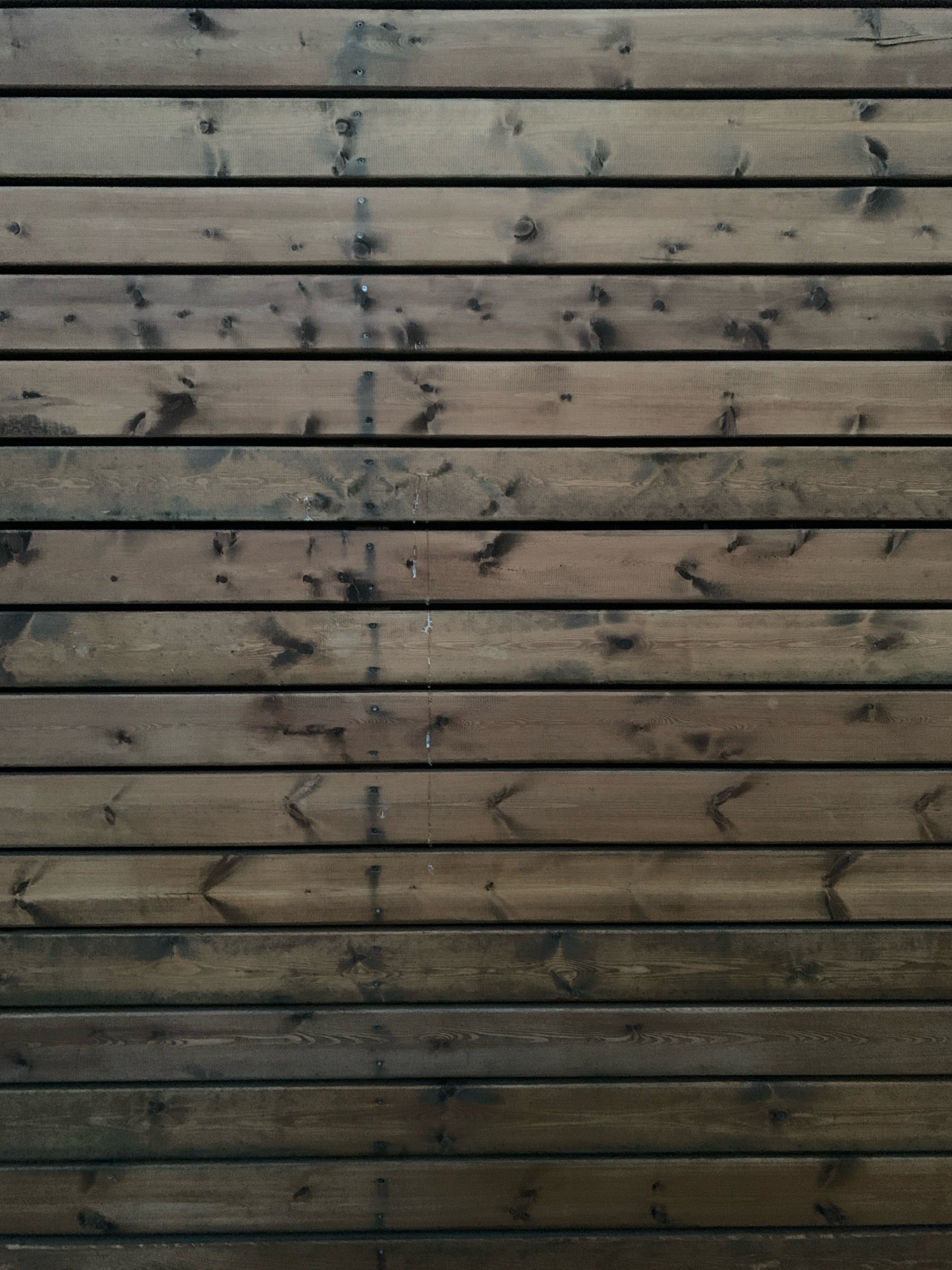 The image shows a close-up, overhead view of a weathered wooden wall in Katunino, Russia. The wall is constructed from horizontal planks, varying in shade from dark brown to a lighter, grayish-brown, with visible wood grain and knots. The planks are uniformly spaced, creating a repeating horizontal pattern. The surface appears to be slightly textured and possibly treated or aged, exhibiting a rustic and natural aesthetic. There are no people or other objects visible in the frame.
