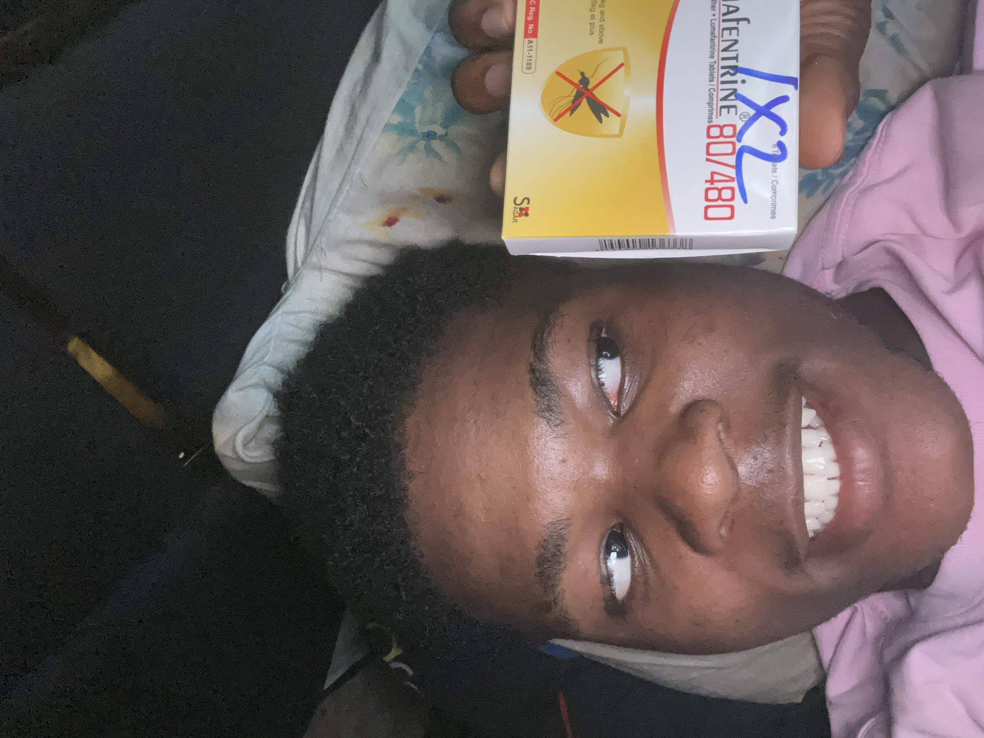 A young Black person is lying in bed, holding a box of malaria medication. The person is looking to their left, with a slight smile on their face. The medication box is yellow and red and has the text "ALAFENTRINE 80/480" and "1X2" written on it in blue ink. The person is wearing a pink t-shirt. The background is dark and out of focus, but appears to be a bedroom. The image was taken in Ihiagwa, Nigeria.