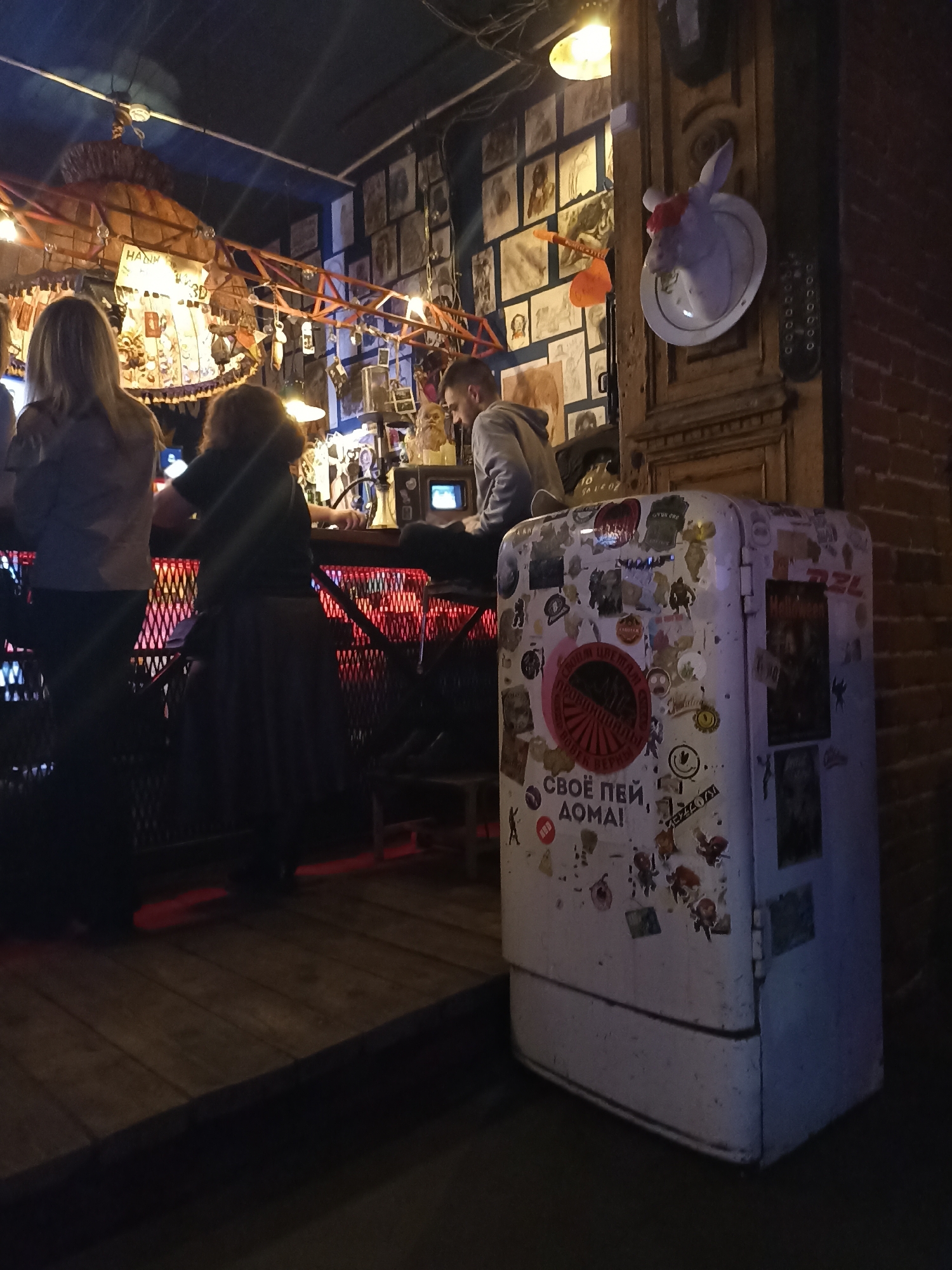 A photo taken in the city of Perm, Russia, depicts the interior of a bar. Three individuals are positioned at the counter; two are women, their backs to the camera, and a man is sitting at the bar. The bar itself is lit by red lights. A vintage-style refrigerator, covered in stickers, stands to the side. On the wall behind the bar, numerous framed images are visible. A decorative deer head is mounted on the wall, positioned on a plate.
