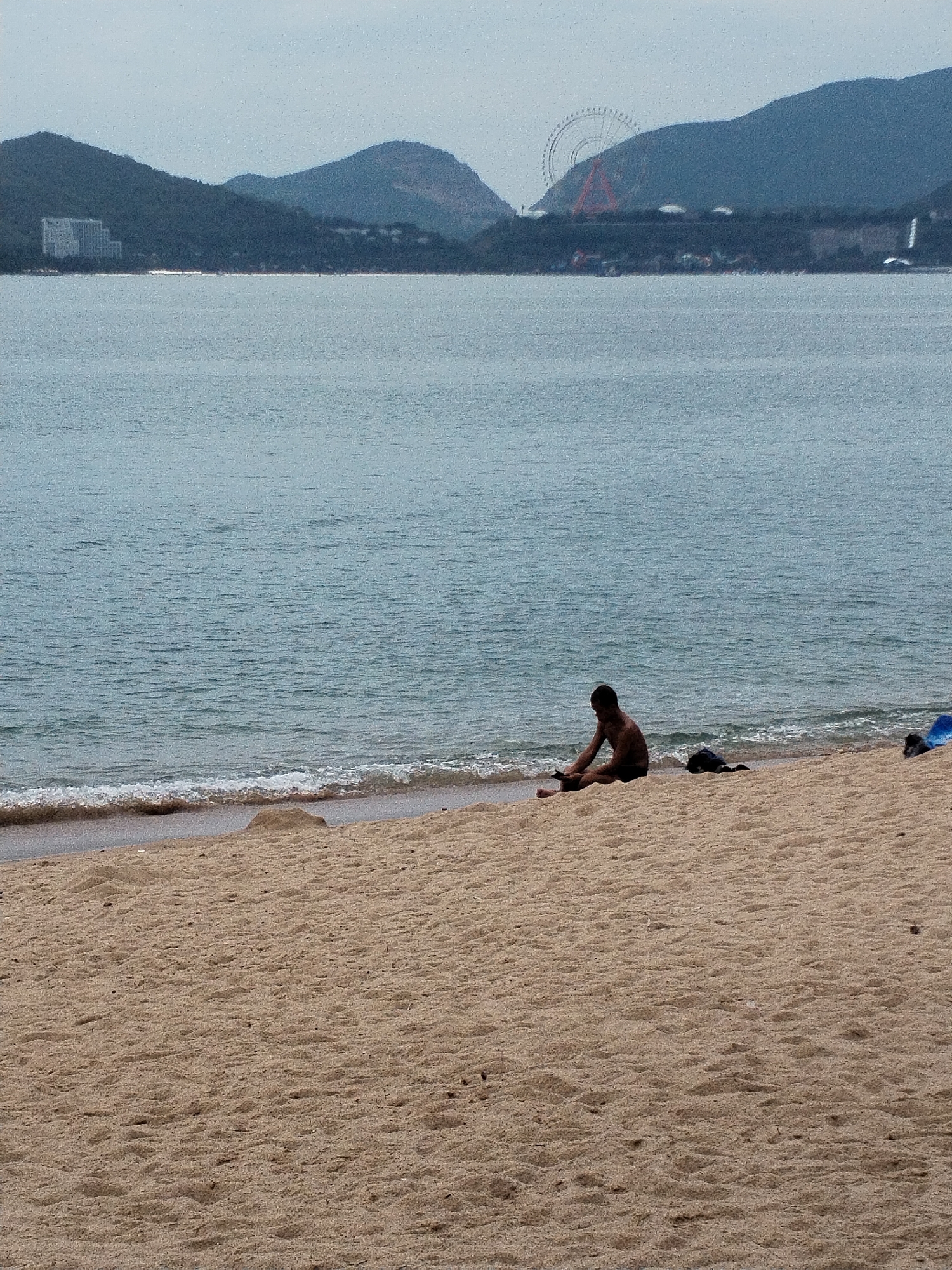 This full-shot photograph captures a tranquil beach scene in Nha Trang Ward, Vietnam, under an overcast sky. The foreground is dominated by light brown sand, showing subtle patterns of footprints and ripples, leading to a gentle shoreline. Small, foamy waves break softly at the water's edge.
In the mid-ground, a lone man, possibly of East Asian descent, is seated directly on the wet sand, facing left and slightly downwards. He appears to be interacting with something in his hands or on the sand in front of him. He is wearing dark swimwear or shorts. To his immediate right, a dark piece of clothing or a small bag rests on the sand, and further to the right, a bright blue object, possibly a towel or another bag, is visible.
The expansive body of water, likely the South China Sea, stretches across the middle of the frame, exhibiting calm, slightly choppy blue-grey hues.
The background features a dramatic mountainous coastline. On the left side of the distant shore, a large, multi-story white building, possibly a hotel or resort, is visible nestled against the green-covered hills. Further to the right, prominently positioned on a distant mountain peak, is a very large Ferris wheel and a distinctive red triangular structure, characteristic of the Vinpearl Land amusement park, aligning with the provided location context of Nha Trang. The mountains themselves are dark green and imposing, contrasting with the lighter tones of the sky and sea.
The overall atmosphere is quiet and serene, suggesting a moment of solitary contemplation or relaxation. No text is visible in the image.