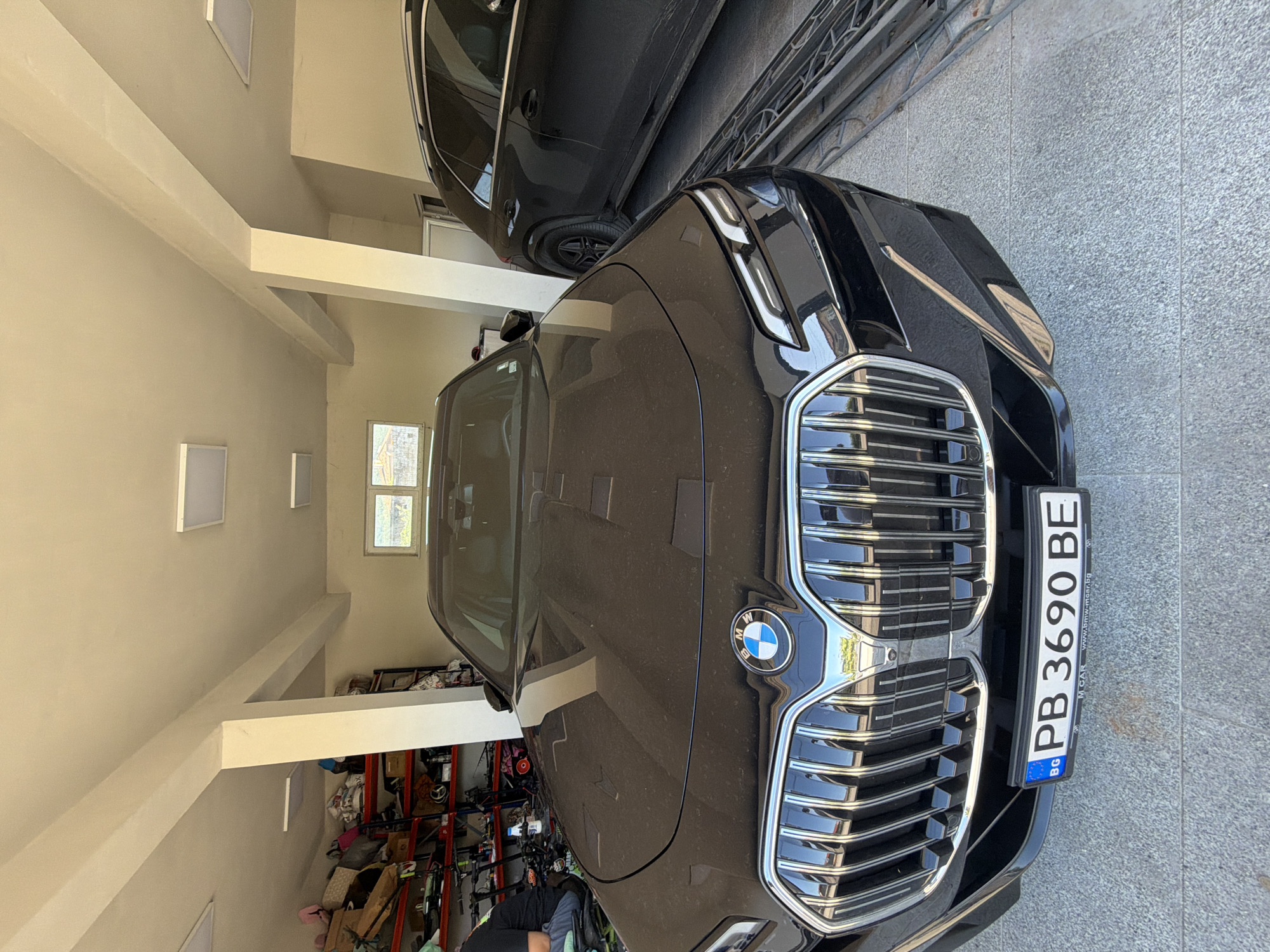 The image shows the front of a black BMW parked in a garage or parking area. The license plate is visible and reads "PB 3690 BE", indicating it is a Bulgarian license plate. The vehicle is parked in front of another black car, partially visible in the top part of the frame. The location appears to be indoors, with a white-painted interior, as evidenced by the ceiling beams and the presence of a window in the background. There is a cluttered storage area behind the vehicle. Given the Bulgarian license plate, the location is possibly in Asenovgrad, Bulgaria, although the specific address or landmark cannot be identified from this single image.