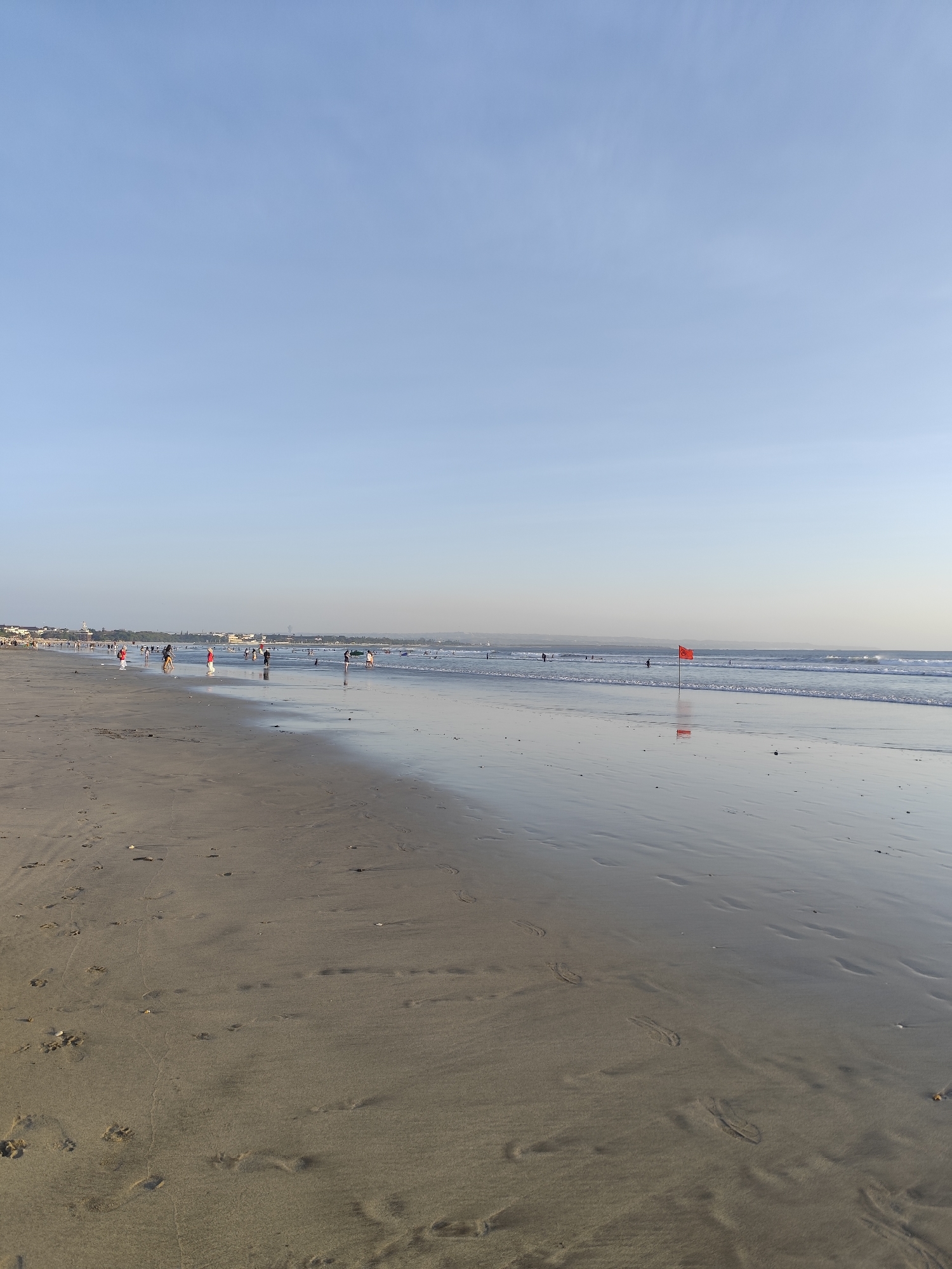 The image depicts a wide, sandy beach with gentle waves lapping the shore. The sky is a clear, pale blue. In the middle distance, a group of people are visible scattered along the shoreline, some walking and others standing near the water. Further out in the ocean, a few individuals appear to be in the water, possibly swimming or surfing. A single red flag is planted on the beach, its reflection visible on the wet sand. The sand itself is marked with numerous footprints. In the far distance, a hazy line of buildings and vegetation suggests a coastal town or resort. The scene appears to be tranquil, with the soft light indicating either early morning or late afternoon.