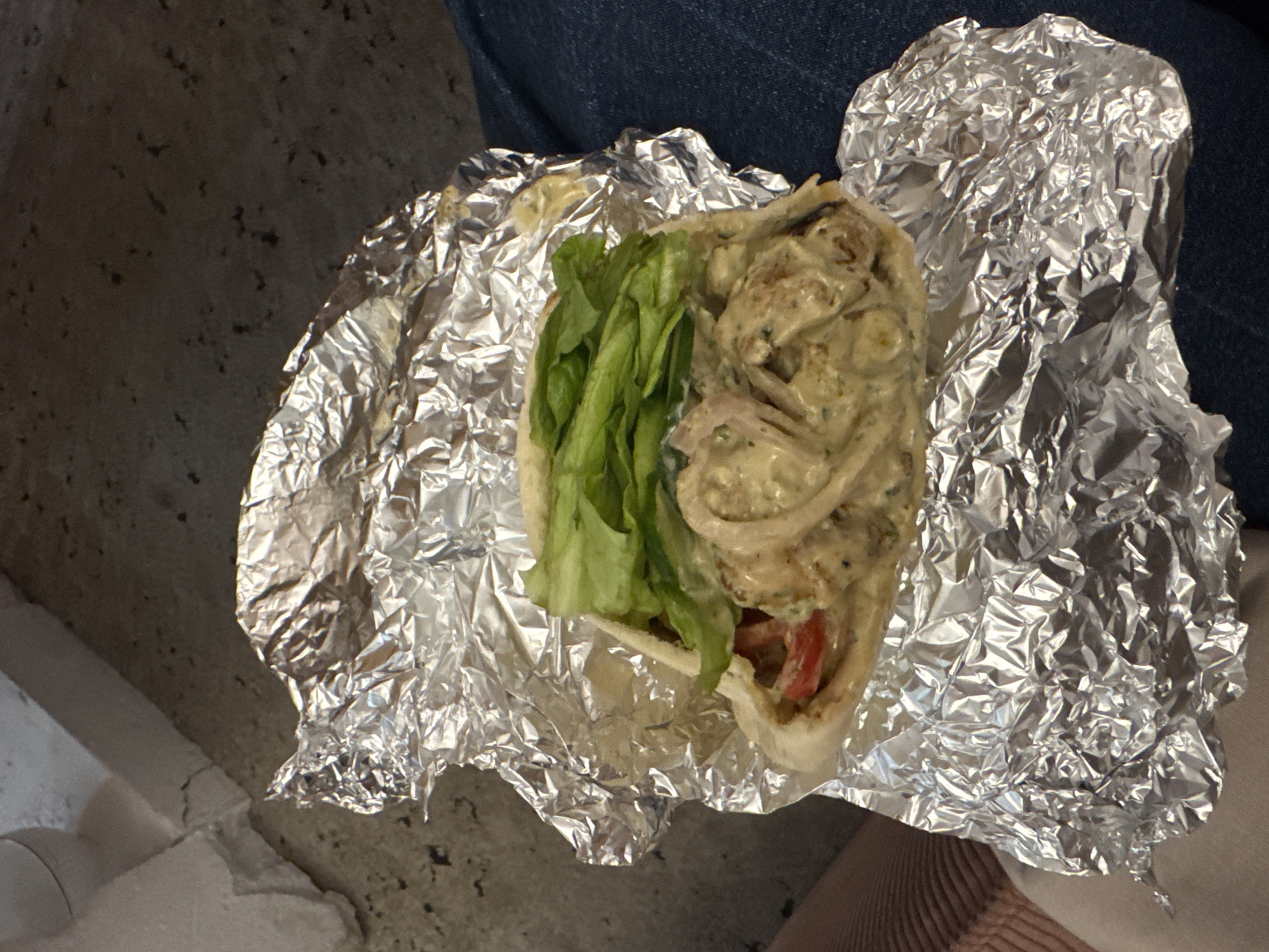 A gyro sandwich wrapped in aluminum foil sits on a person's lap. The sandwich contains lettuce, tomato, and a creamy meat filling. The person is wearing jeans. The location is unknown.