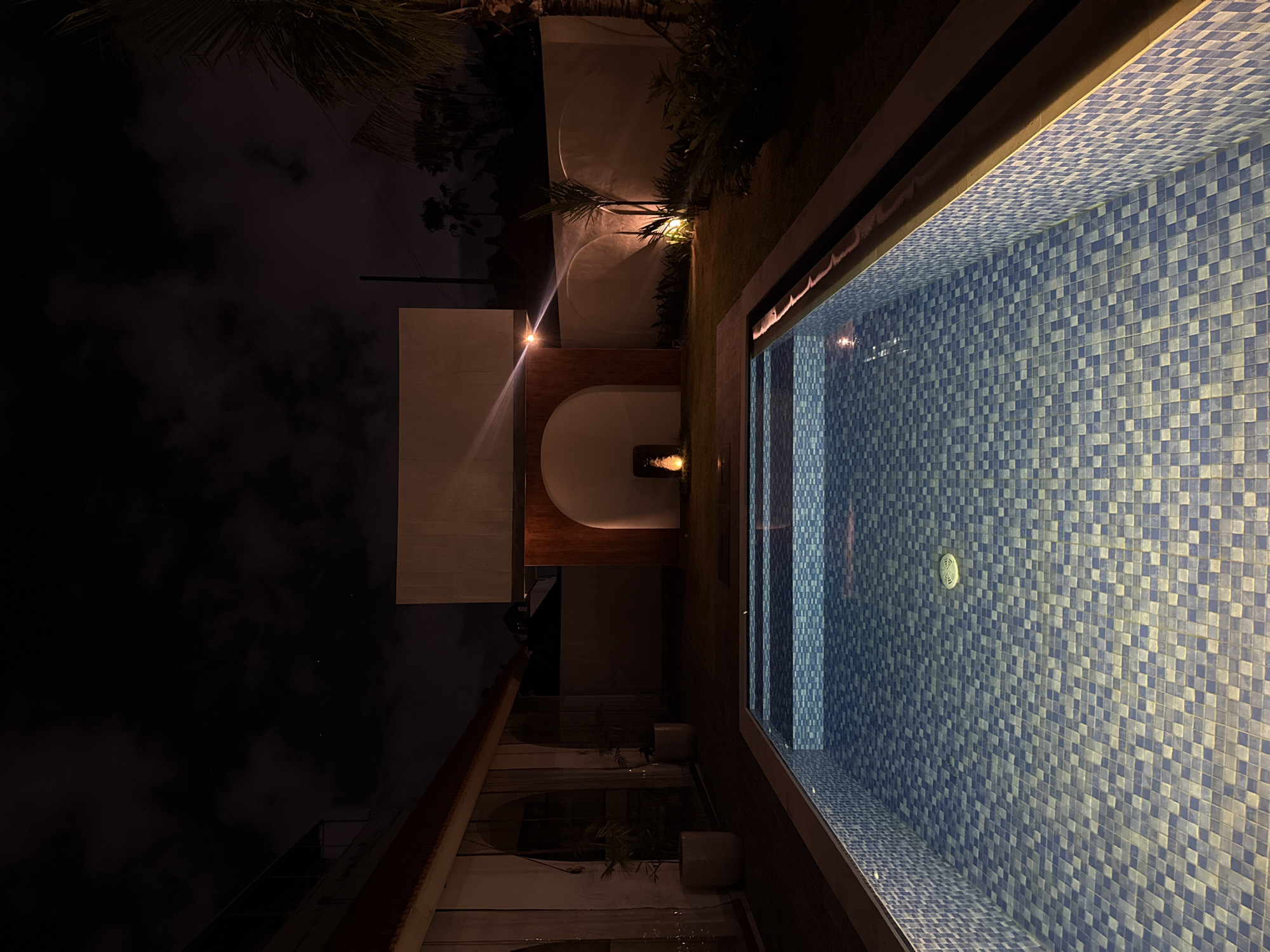 The image captures a serene nighttime view of an outdoor area, likely a luxurious resort or private villa in North Kuta, Indonesia. The scene is dominated by a swimming pool on the right and an illuminated architectural and landscape feature on the left.
The swimming pool features striking blue and white mosaic tiles, arranged in a checkerboard pattern, covering its interior. The water is clear, reflecting the artificial lighting from within and around the pool, creating a gentle shimmer. A circular drain or inlet fixture is visible on the upper part of the pool wall. The pool deck or edge is dark-toned, possibly wood or stone, contrasting with the bright tiles.
To the left, a modern building or wall structure is partially visible, featuring a large, illuminated arched cutout. Inside this arch, a warm light source highlights a decorative element, possibly a small fountain or sculpture. Another bright light source is positioned on the wall to the left of the arch, casting a strong glare and illuminating nearby tropical foliage. The surrounding area includes lush green grass and several palm trees, partially silhouetted against the dark night sky. Some clouds are discernible in the sky, adding texture to the otherwise dark expanse. The artificial lighting creates a warm and inviting atmosphere, enhancing the tranquil ambiance of the outdoor space. No people are visible, and no specific activity is taking place. There is no visible text.