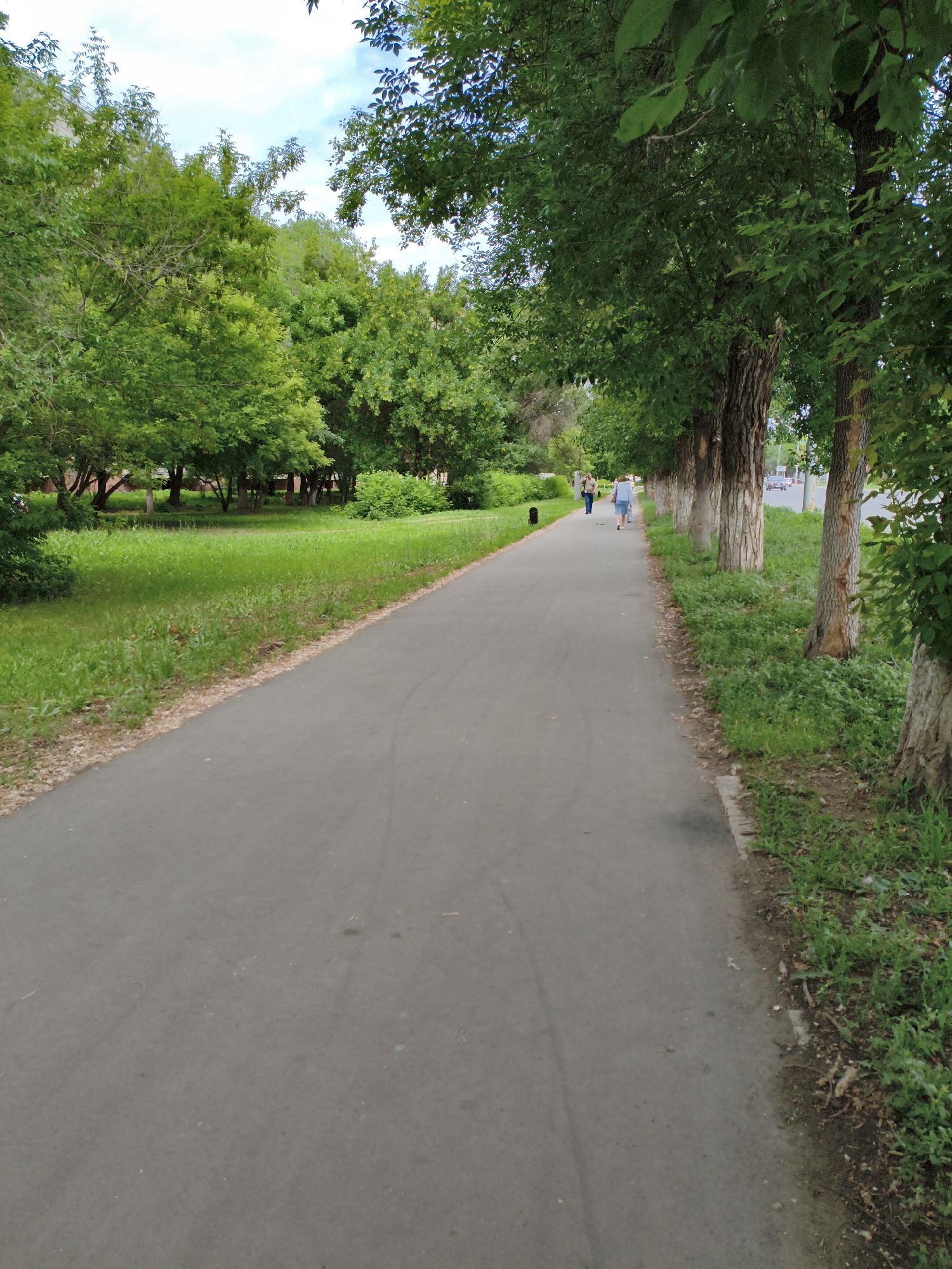 The image depicts a paved pathway in a park-like setting, likely in Orenburg, Russia. The pathway is bordered by lush green grass and a row of mature trees with thick, textured bark. On the right side of the path, the trees form a dense canopy, casting shadows. On the left side of the path, the greenery extends into a mown lawn with scattered bushes. In the distance, two individuals are walking away from the camera along the pathway. One appears to be wearing a light-colored top and darker bottoms, while the other is in a light blue top and darker bottoms. Further in the distance, beyond the park area, a road with passing vehicles is visible. The sky is partly cloudy with patches of blue. The overall scene suggests a pleasant day in an urban park.