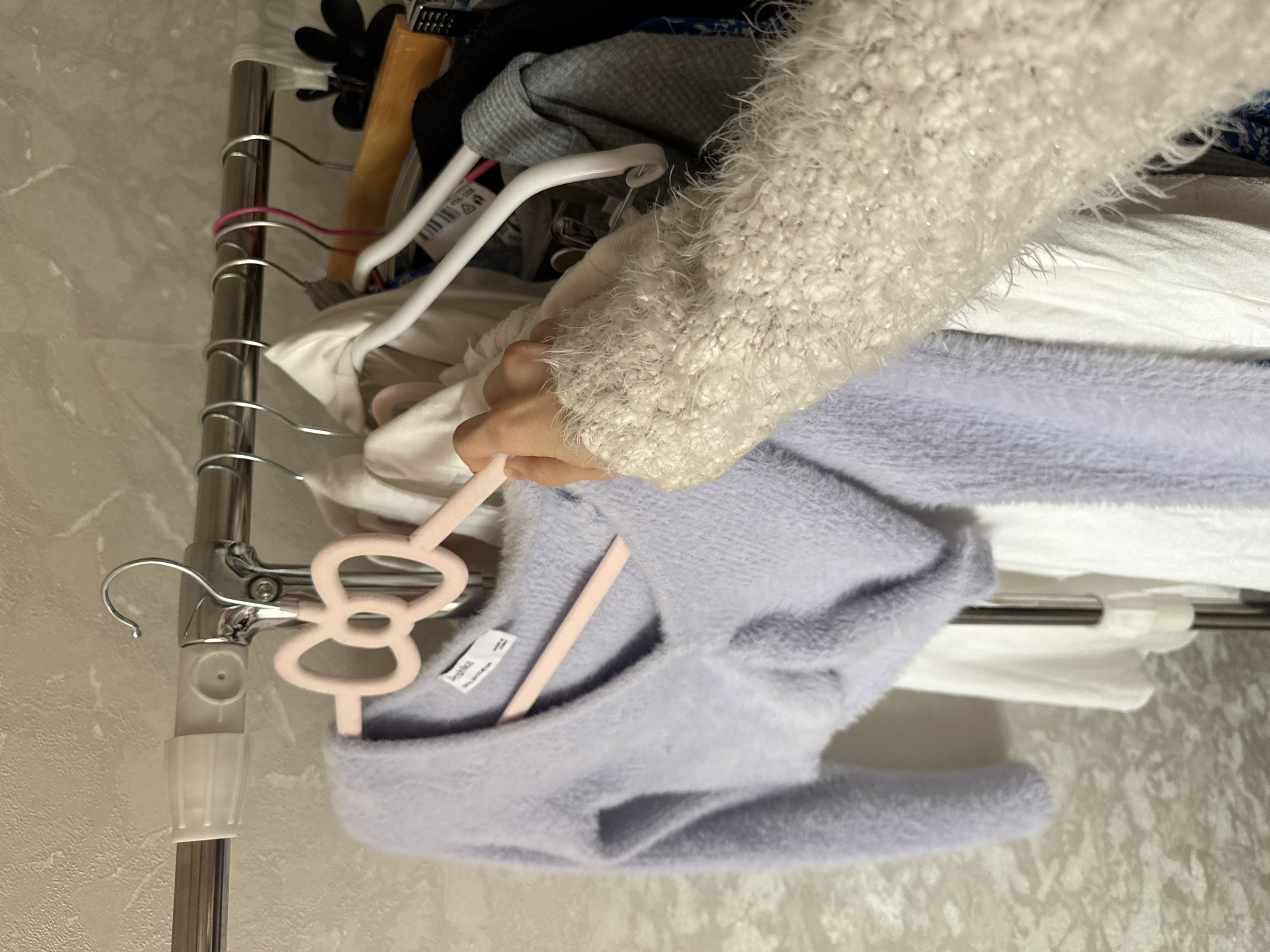 A hand, wearing a fuzzy white sweater, reaches out to adjust a light purple fuzzy sweater hanging on a pink bow-shaped hanger. The purple sweater is on a clothing rack next to other garments, including white shirts and a grey top. The background is a textured, light-colored wall. The scene appears to be an indoor closet or dressing area.