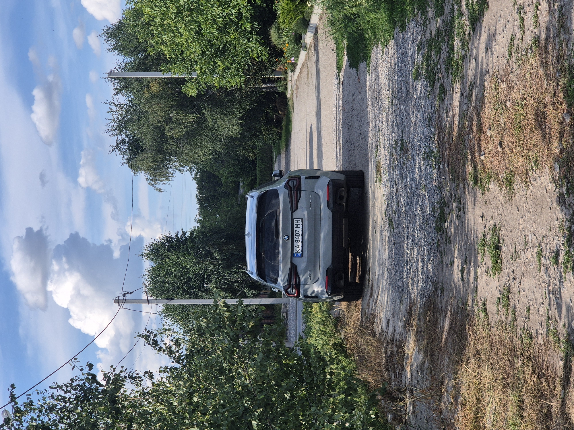 A light-colored BMW SUV is parked on a gravel road surrounded by trees and greenery. The car is viewed from the rear, displaying its license plate which reads "KA 8407 MH". The road is partially covered with small stones and dirt. Overhead, a blue sky with fluffy white clouds is visible, with utility poles and wires present in the background. The scene appears to be in a rural or semi-rural area, consistent with the village of Sozonivka, Ukraine. The overall impression is a quiet, daytime scene in an outdoor setting.