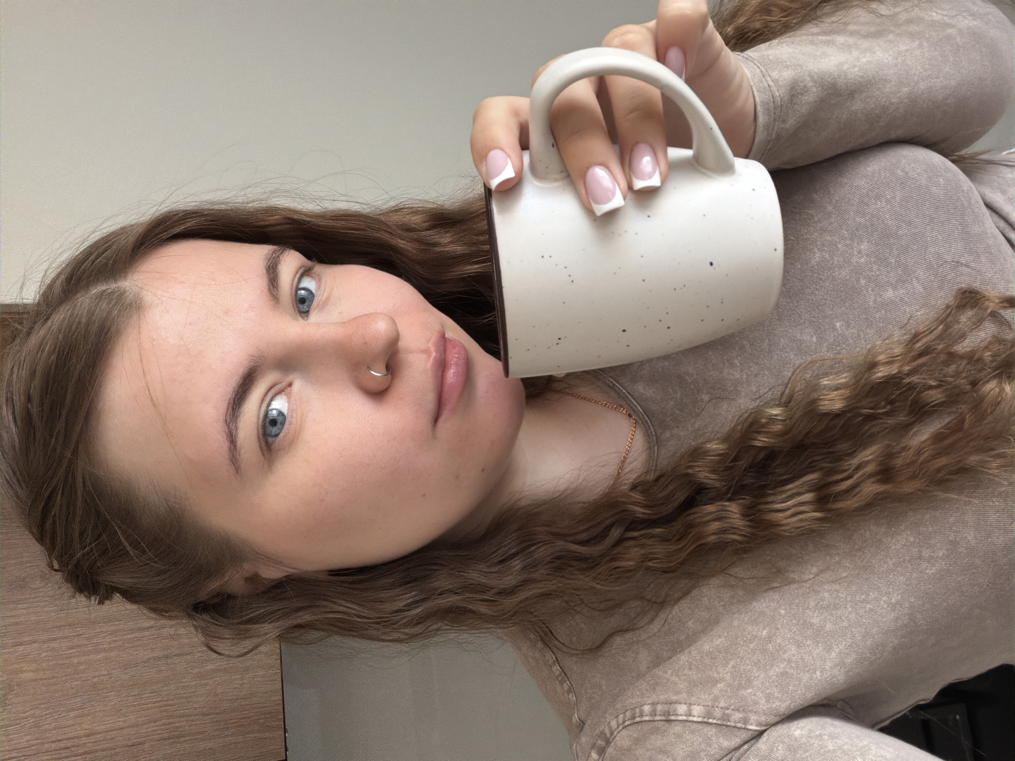 A woman with fair skin, blue eyes, and light brown, curly hair holds a white mug with black speckles in her right hand. She wears a long-sleeved, light brown shirt and a delicate gold necklace. She has a silver ring in her left nostril and neatly manicured nails with white tips. The background is a plain, light gray wall above and a wooden-look surface below. There's no indication of the location being Chaikovsky, Russia.