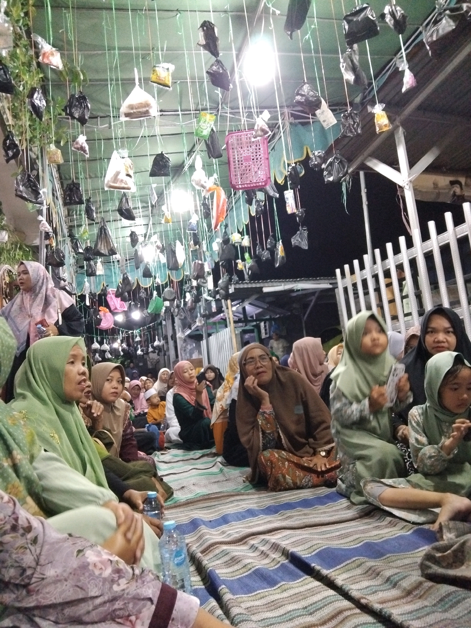 This image depicts an outdoor gathering at night in Kepuh Kiriman, Indonesia. The scene is characterized by an overhead canopy strung with numerous small plastic bags of various colors and contents, suspended by thin cords. A group of people, predominantly women and children, are seated on the ground, which is covered with striped mats. Most of the individuals are wearing traditional Islamic attire, including hijabs and modest clothing in muted tones. Some individuals appear to be engaged in conversation, while others are looking in various directions. The lighting in the scene is dim, with artificial lights visible overhead, possibly illuminating the hanging bags and creating reflections. In the background, there is a white fence structure and a dark, outdoor environment. The overall atmosphere suggests a communal event or gathering taking place in a residential or public outdoor space.