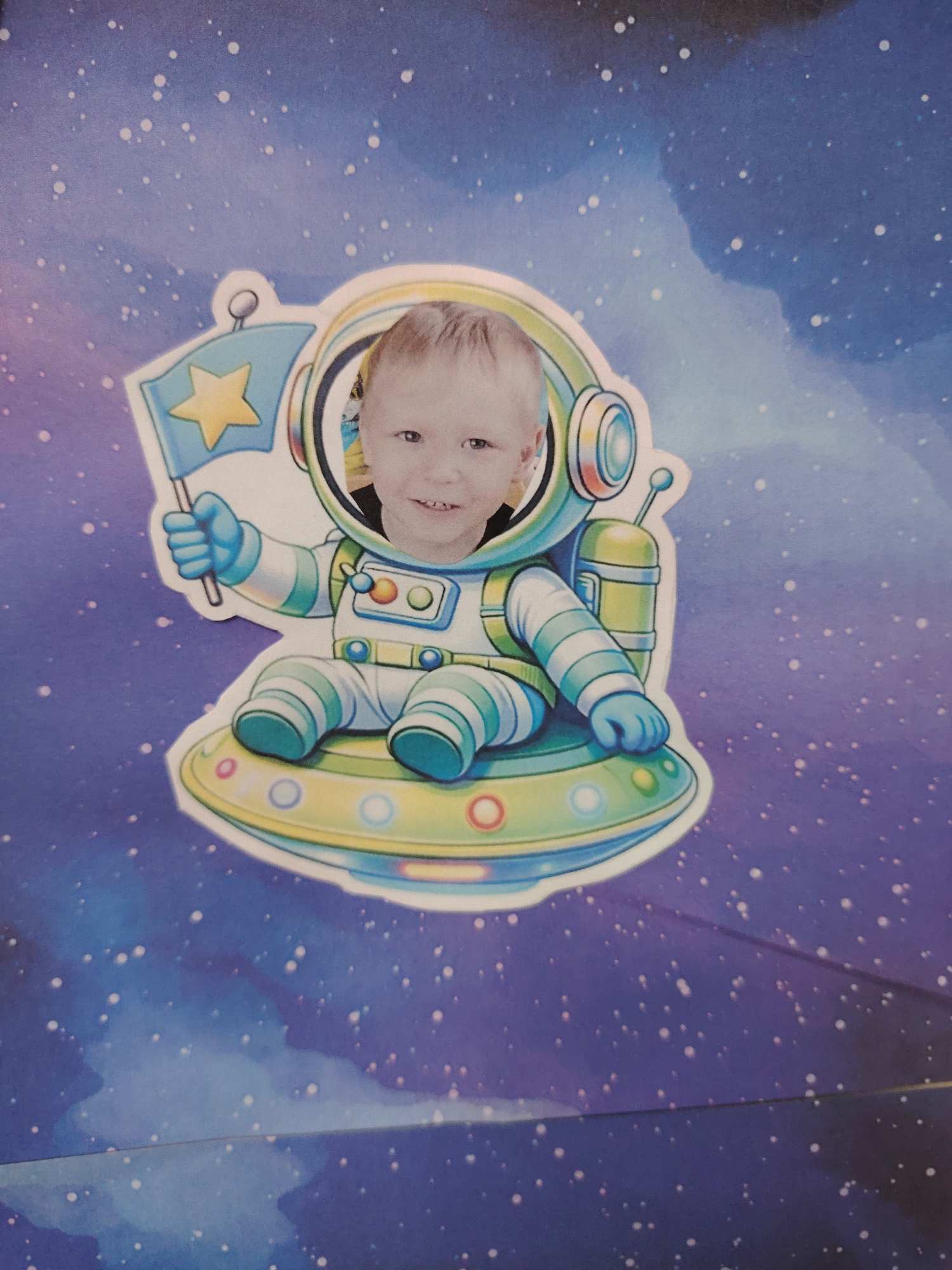 A close-up photograph shows a child's image superimposed on a cartoon astronaut sitting in a flying saucer, against a backdrop of a space-themed design. The astronaut is holding a flag with a star. The location of the image, Sarapul, Russia, is not discernible from the visual content.