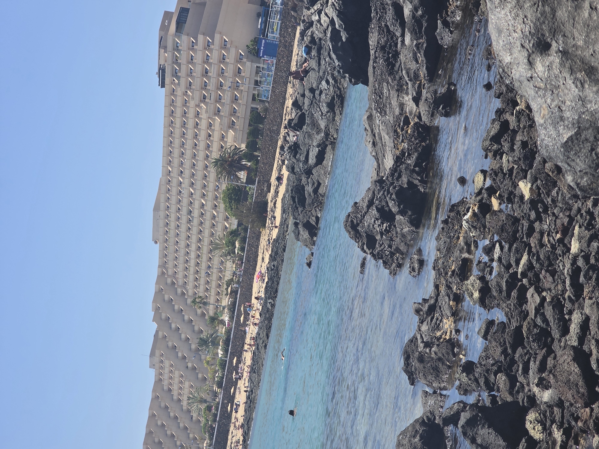 The image depicts a coastal scene in Costa Teguise, Spain. In the foreground, dark volcanic rocks are partially submerged in clear turquoise water. Further back, a sandy beach is visible, populated with numerous sunbathers relaxing under yellow umbrellas. Behind the beach, a large, multi-story hotel complex dominates the landscape, its tiered architecture creating a distinctive zigzag pattern. Palm trees are interspersed between the hotel and the beach. The sky is clear and blue. In the water, two individuals are present; one is swimming, and another appears to be partially submerged, possibly also swimming or wading. The overall impression is a sunny day at a popular tourist resort.