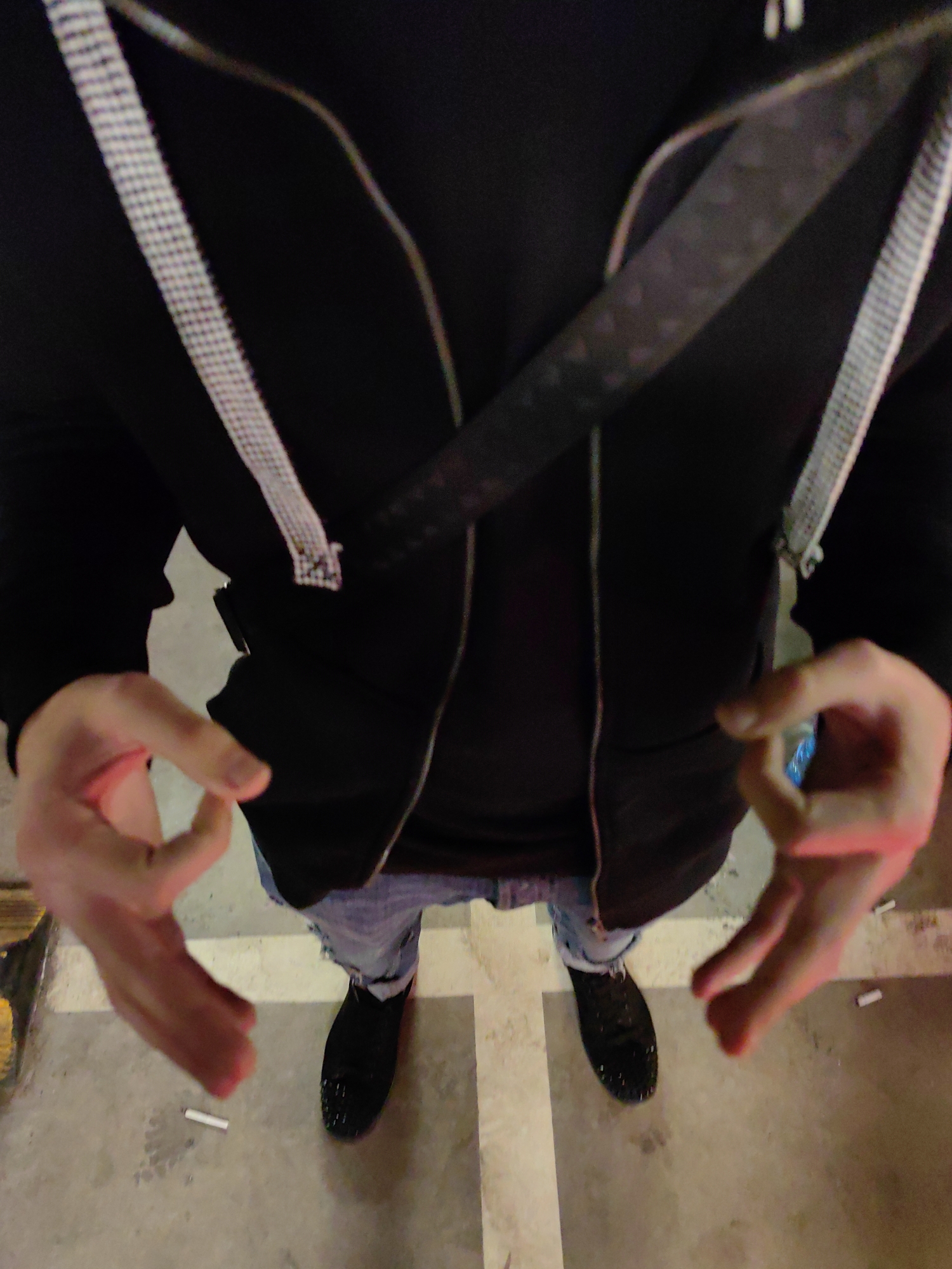A person stands in what appears to be a parking garage, judging by the marked parking space on the floor. The person is wearing black shoes, light blue jeans, a black t-shirt, and a black hooded jacket. A strap is worn across the chest. The person's hands are raised in the air, making "OK" gestures. Discarded cigarette butts are visible on the ground. The specific location within Sofia, Bulgaria, cannot be determined from the image.