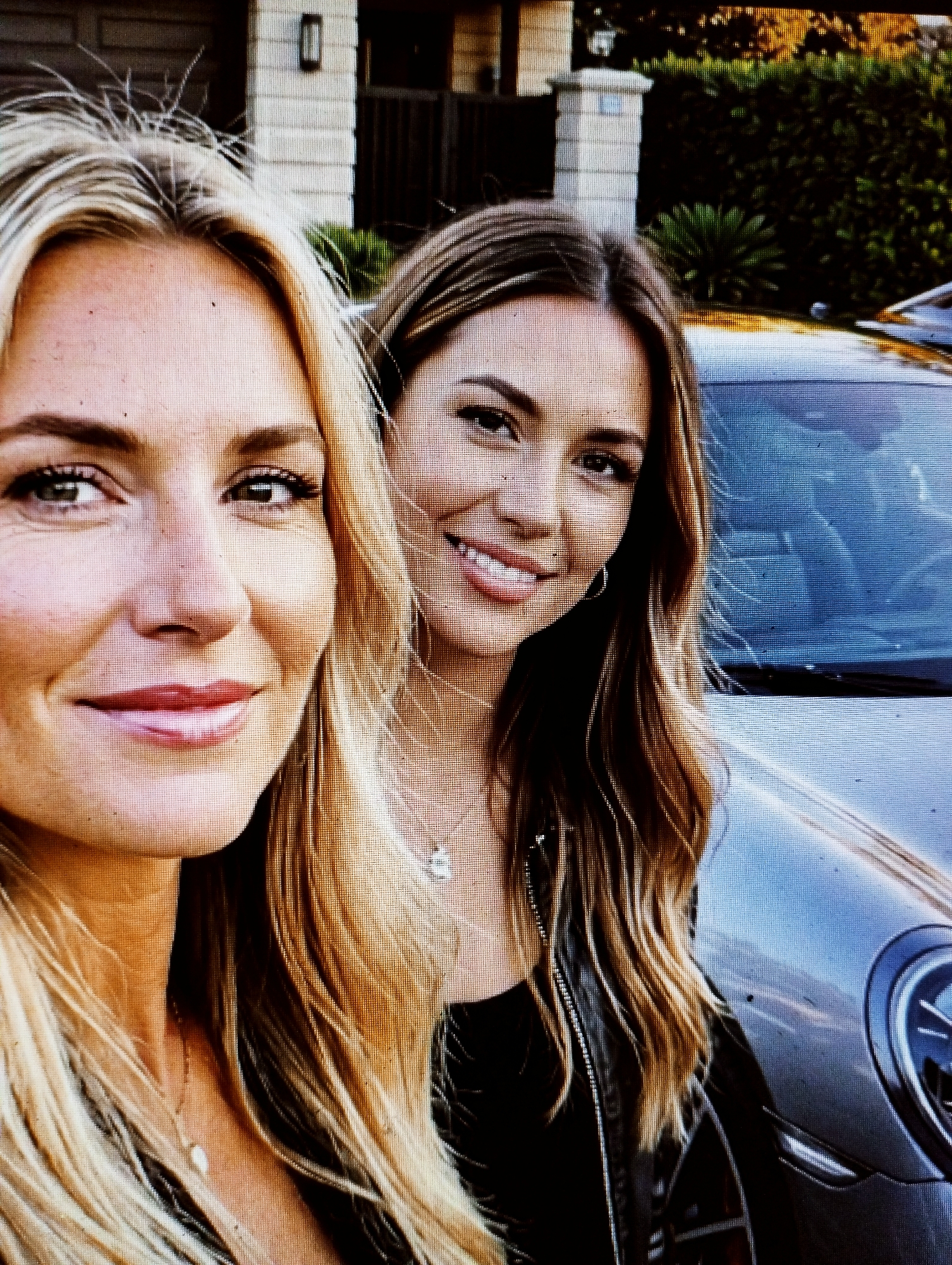 Two women are posing for a selfie outdoors. The woman on the left is in the foreground, with her face close to the camera. She has blonde hair and is smiling. She is wearing a necklace. The woman on the right is partially visible behind her. She has brown hair and is also smiling. She is wearing a black jacket and a necklace with a small pendant. They are standing in front of a silver car. In the background, there is a building with light-colored siding and a dark roof, and some green foliage. The lighting suggests it is daytime.