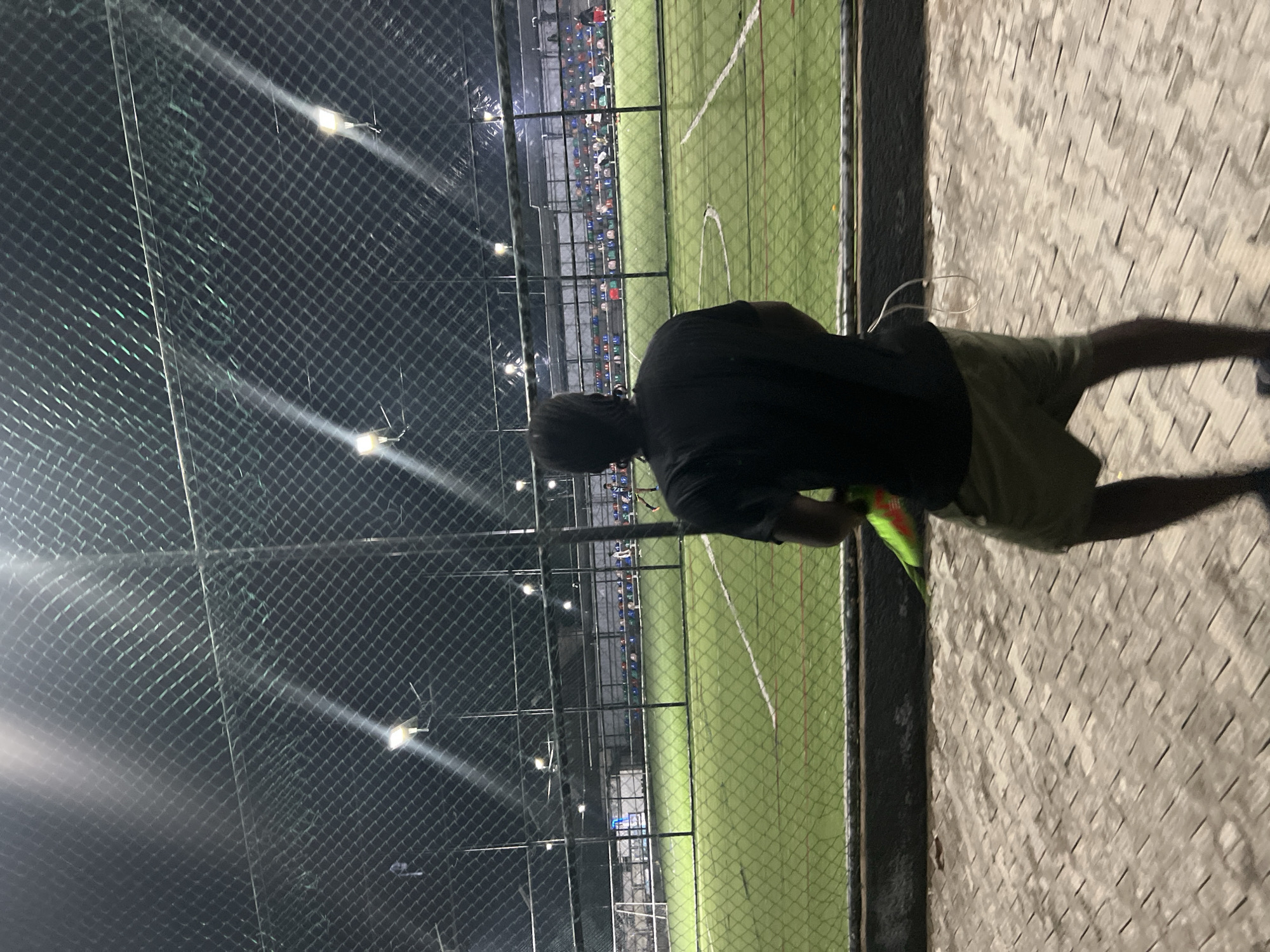 An individual stands facing away from the camera, looking towards a brightly lit artificial turf soccer field at night. The field is enclosed by a tall chain-link fence and features white markings for the game. In the background, tiered seating filled with spectators is visible. The individual is wearing a black t-shirt and khaki shorts and is holding a bright green and orange object. The foreground consists of a patterned paved walkway. The scene is located at a sports facility in Jalingo, Nigeria.