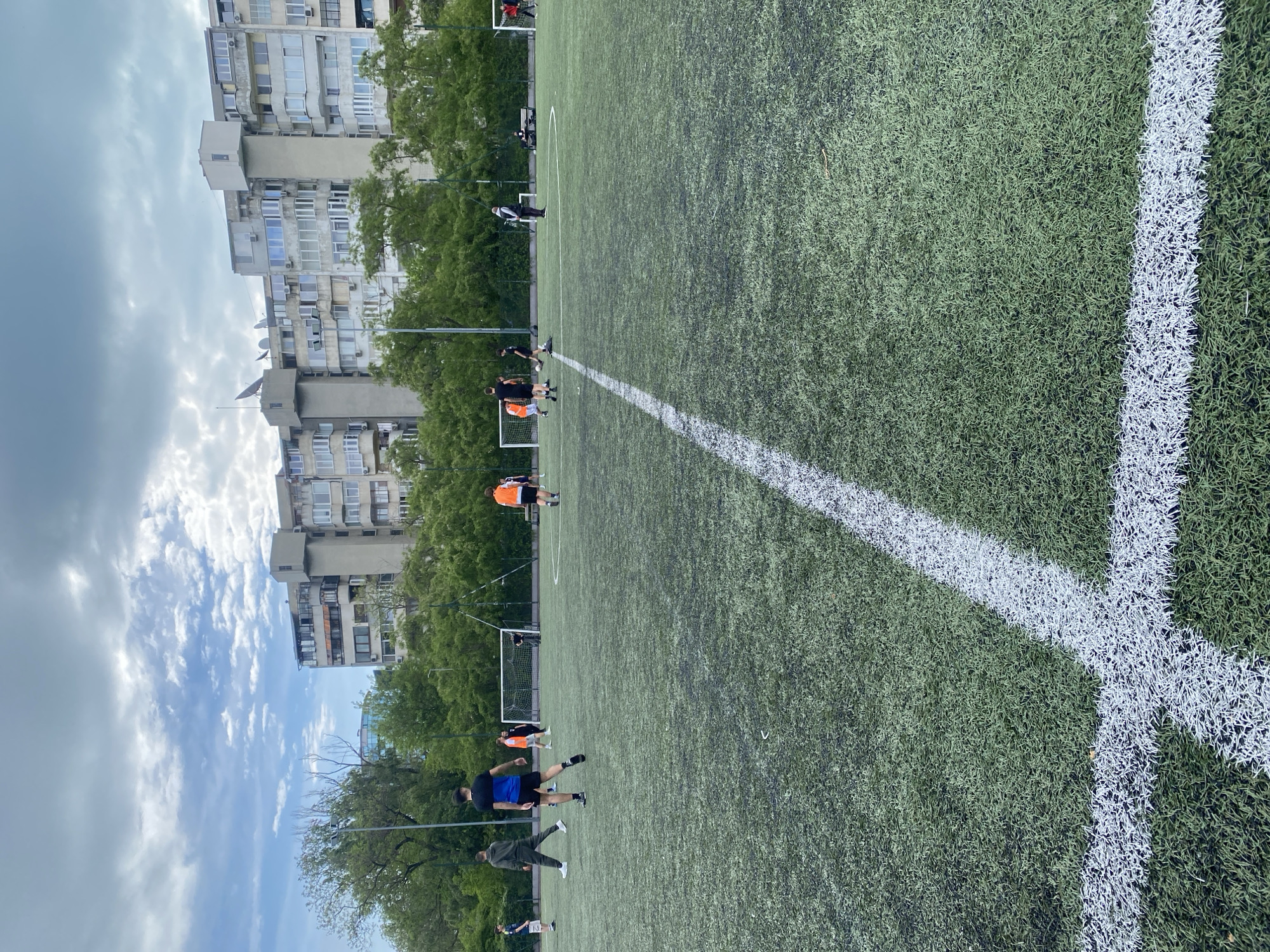 The image depicts a soccer field with artificial turf in Varna, Bulgaria. Players are scattered across the field, some wearing orange and black uniforms, others in blue. The field is marked with white lines. Behind the field, apartment buildings rise into a cloudy sky. Trees are interspersed between the field and the buildings. A soccer goal is visible at the far end of the field.