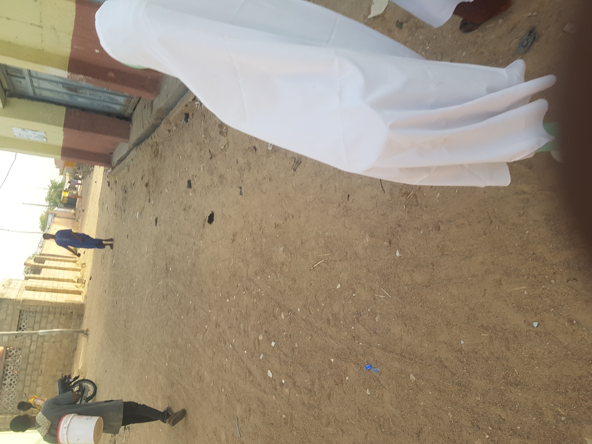 The image depicts a dirt road in Dabawa, Nigeria. Buildings line the left side of the road. Two individuals are visible on the left side. One is on a motorbike and carries a white bucket, and the other is wearing blue. A large white sheet fills the upper right quadrant of the image.