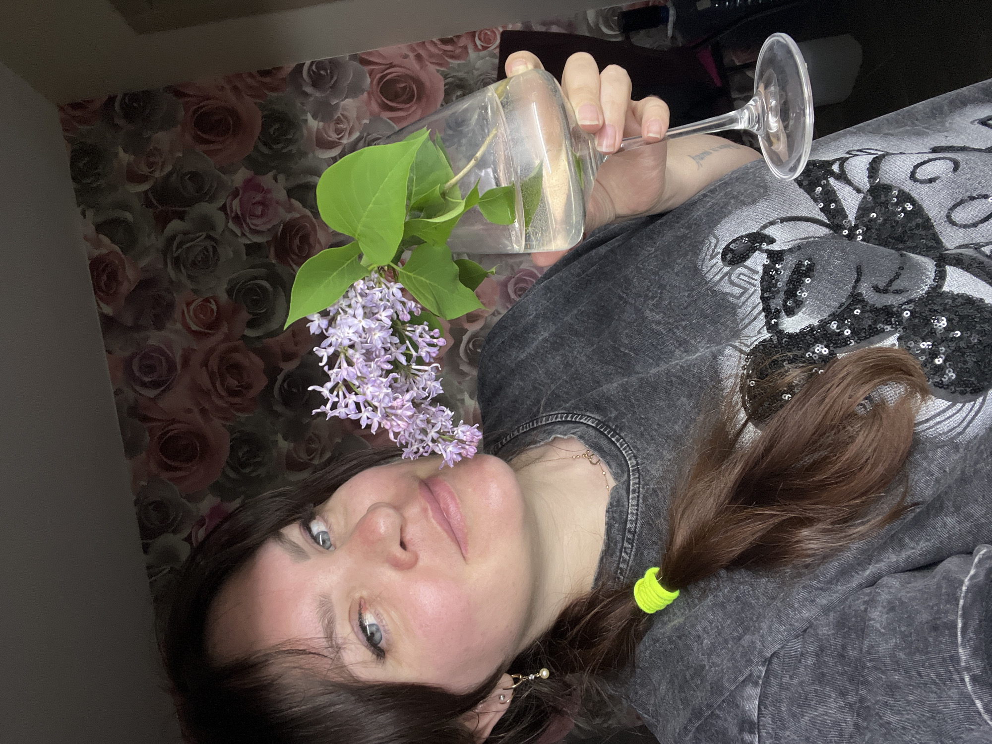 A person is holding a wine glass with lilac flowers in it. The person is wearing a gray shirt and has brown hair pulled back with a green scrunchie. The background is a rose-patterned wallpaper. The person appears to be indoors. The location in Noviy, Russia cannot be determined from the image.