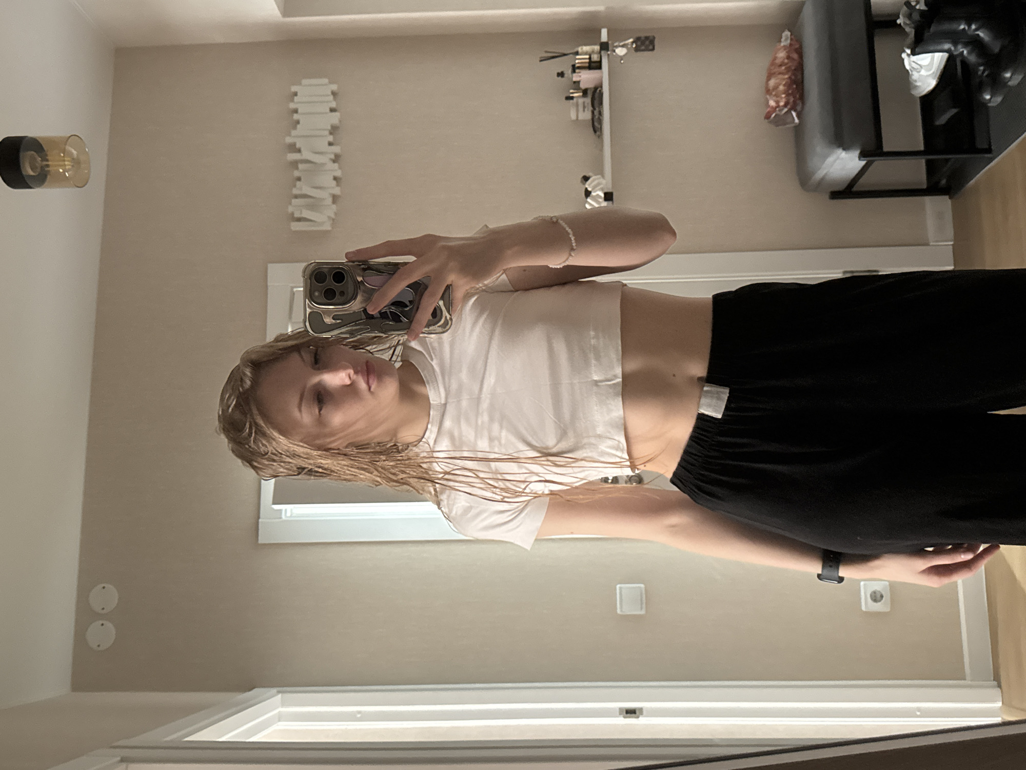 A young woman with wet blonde hair is taking a selfie in a mirror. She is wearing a white crop top and black sweatpants. A smartwatch is on her left wrist. Her phone is a light color.
The environment appears to be a bathroom. The walls are a pale beige. There's a light fixture on the left wall, a white decorative piece, and shelving on the top right. In the background, there is a bench or seat and some shoes. A window is visible partially behind the woman.
It is not possible to determine if this image was taken in Perm, Russia with the given information.