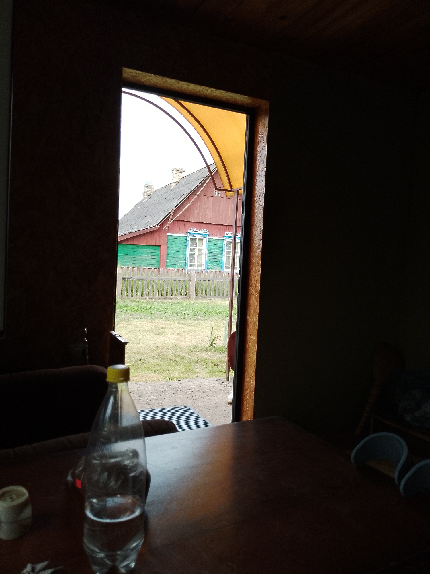 A low-angle, medium shot shows a plastic bottle with a yellow cap sitting on a wooden table in the foreground. The bottle contains a clear liquid and appears to be partially full. Behind the bottle, a large doorway frames a view of a rural scene. To the left, a dark brown wall occupies the foreground. The doorway reveals a red wooden house with a green horizontal siding on its lower portion. The house has a gray roof and a chimney. A wooden fence stands in front of the house, and a grassy yard surrounds it. The sky is overcast. The lighting is dim, with light filtering in from the doorway. The image is likely taken in Bahushewsk, Belarus, given the rural setting and architectural style.