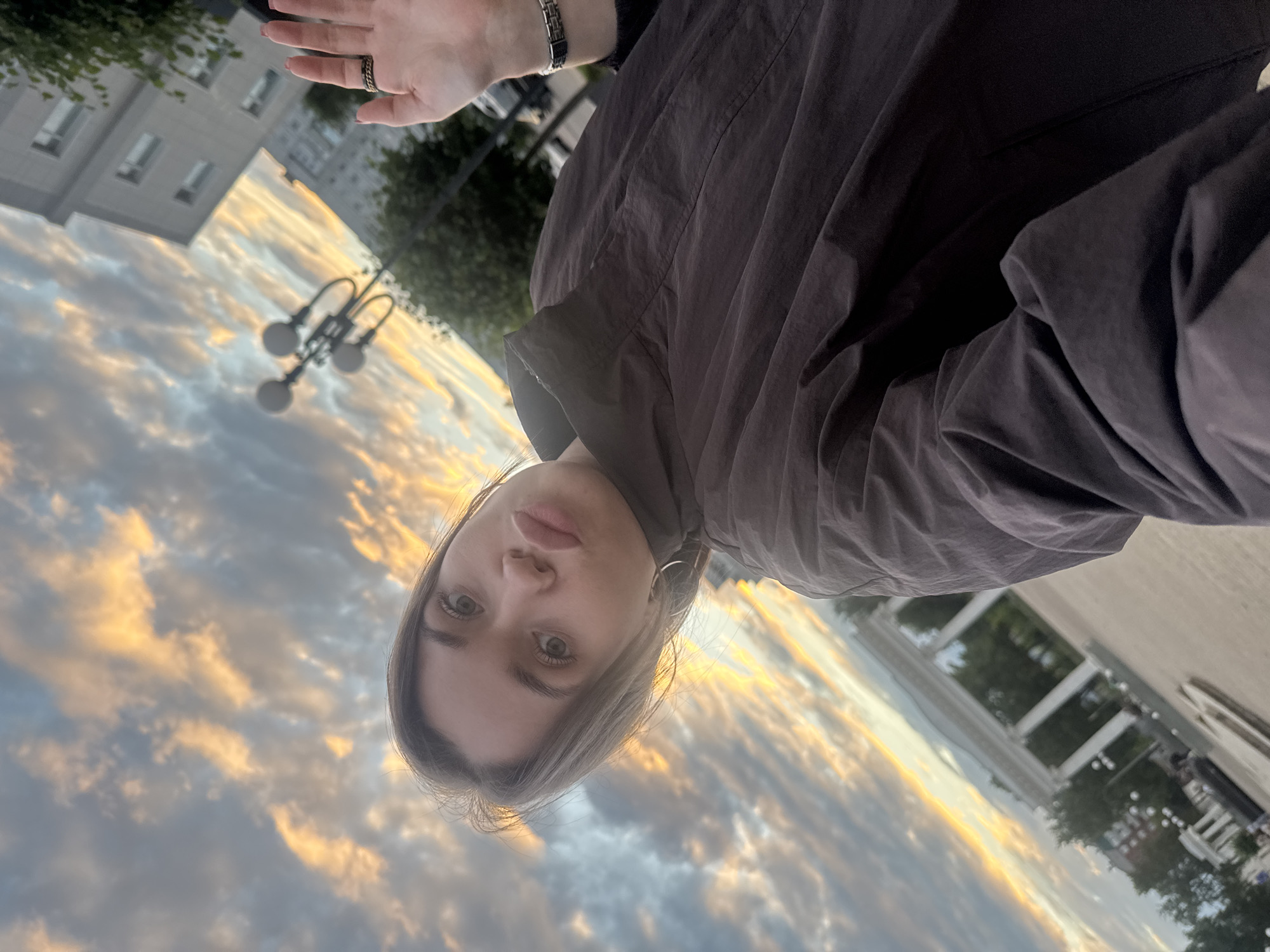 A young woman with blonde hair, wearing a dark jacket, poses for a selfie outdoors against a backdrop of a dramatic sunset sky with clouds in shades of orange and grey. Her mouth is pursed in a kiss-like expression. In the background, to the right, is a modern building and trees. To the left, there is an arched structure with columns, possibly part of an architectural landmark or park. The overall scene suggests an urban setting during twilight. The location is the city of Chaikovsky, Russia, as indicated by the distinctive architectural elements and the general atmosphere.