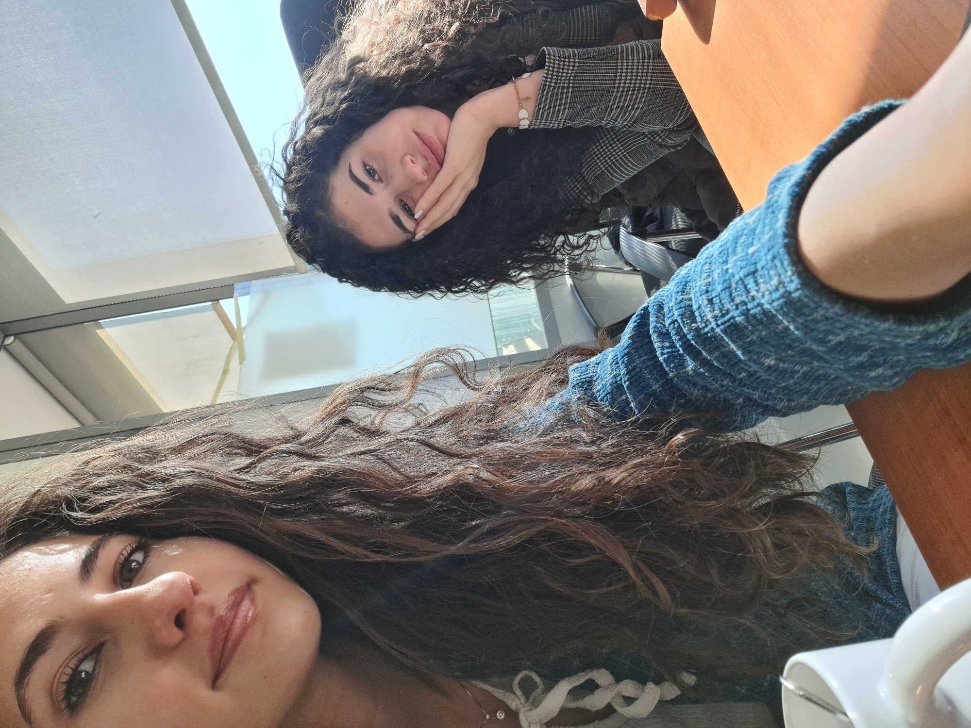 Two young women are posing for a selfie. One woman, with long dark curly hair and wearing a blue sweater, is in the foreground. The second woman, also with dark curly hair, is wearing a gray patterned blazer and resting her face on her hand. They are sitting at a wooden table. A white mug is partially visible in the lower right corner. The background shows a window with white blinds and a view of a bright, sunny sky. Given the architectural details visible through the window, it is impossible to specifically identify the location in Sofia, Bulgaria.