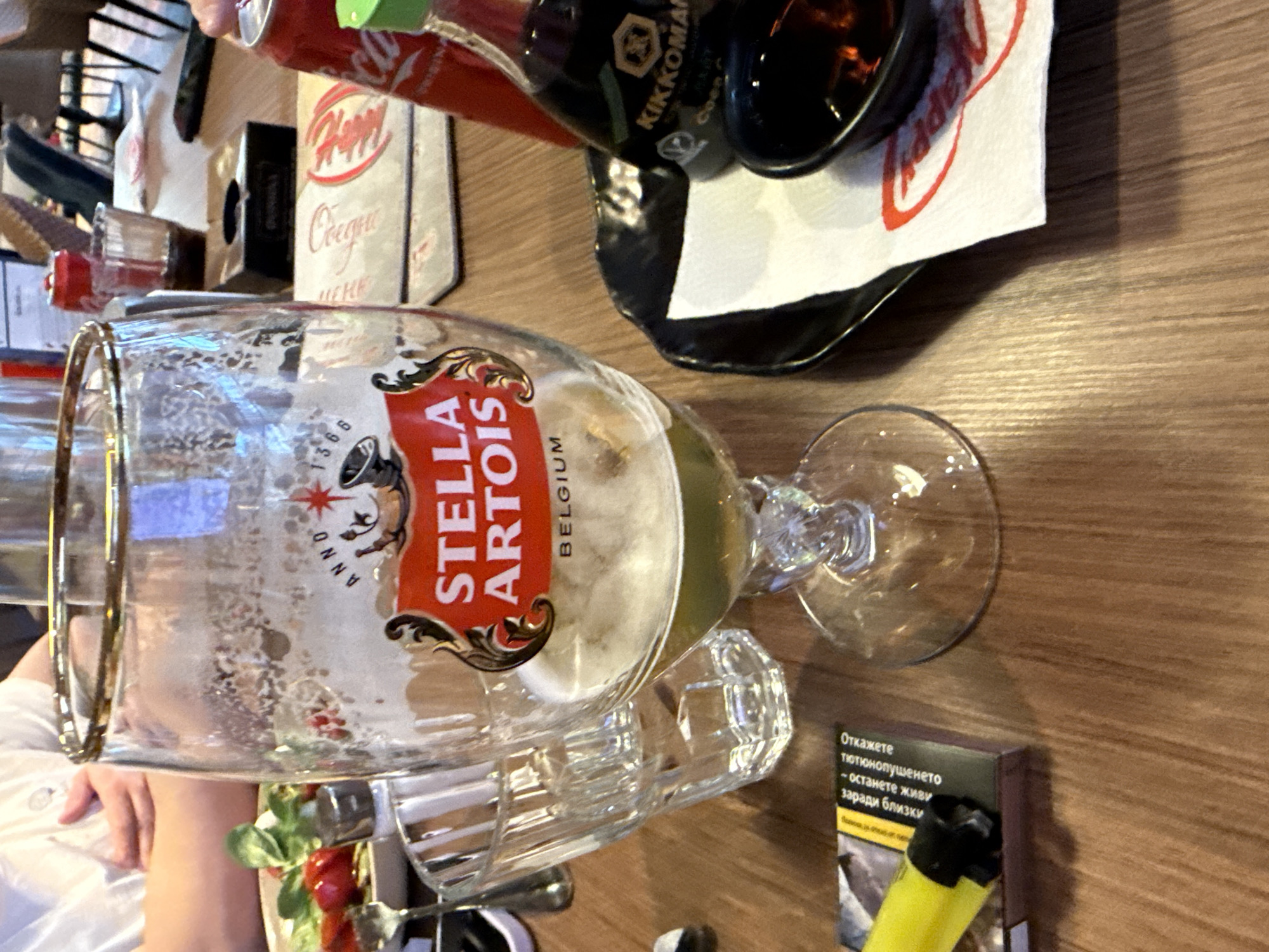 The image shows a table setting in what appears to be a restaurant or bar in Sofia, Bulgaria. A half-filled Stella Artois beer glass is prominently displayed. Next to it is a pack of cigarettes with a yellow lighter, and other items are partially visible, including a napkin with a "Happy" logo, a bottle of Kikkoman soy sauce, and possibly other condiments or food items. A person's arm wearing a white shirt is visible on the left side of the image. The setting suggests a casual dining or social environment.