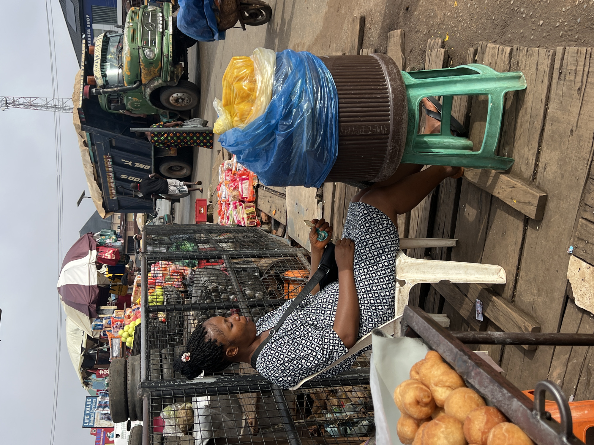 In Nkpor, Nigeria, a woman sits on a white plastic chair next to a large brown barrel covered with blue and yellow plastic bags. She wears a black and white patterned dress and has her hair braided in an updo. In her hand, she holds a small item, possibly food. In front of her, on a metal stand, are several fried dough pastries. To her right, a green plastic stool supports the barrel. The scene is set on a wooden platform, suggesting an outdoor market or roadside stall. In the background, a large green truck with the words "CHUKWUEBUKA INVEST" is parked, and various market stalls with umbrellas are visible, along with stacks of tires and packaged goods. Several other people are present in the background, moving around the market. The sky is overcast, and the overall impression is that of a busy commercial area.