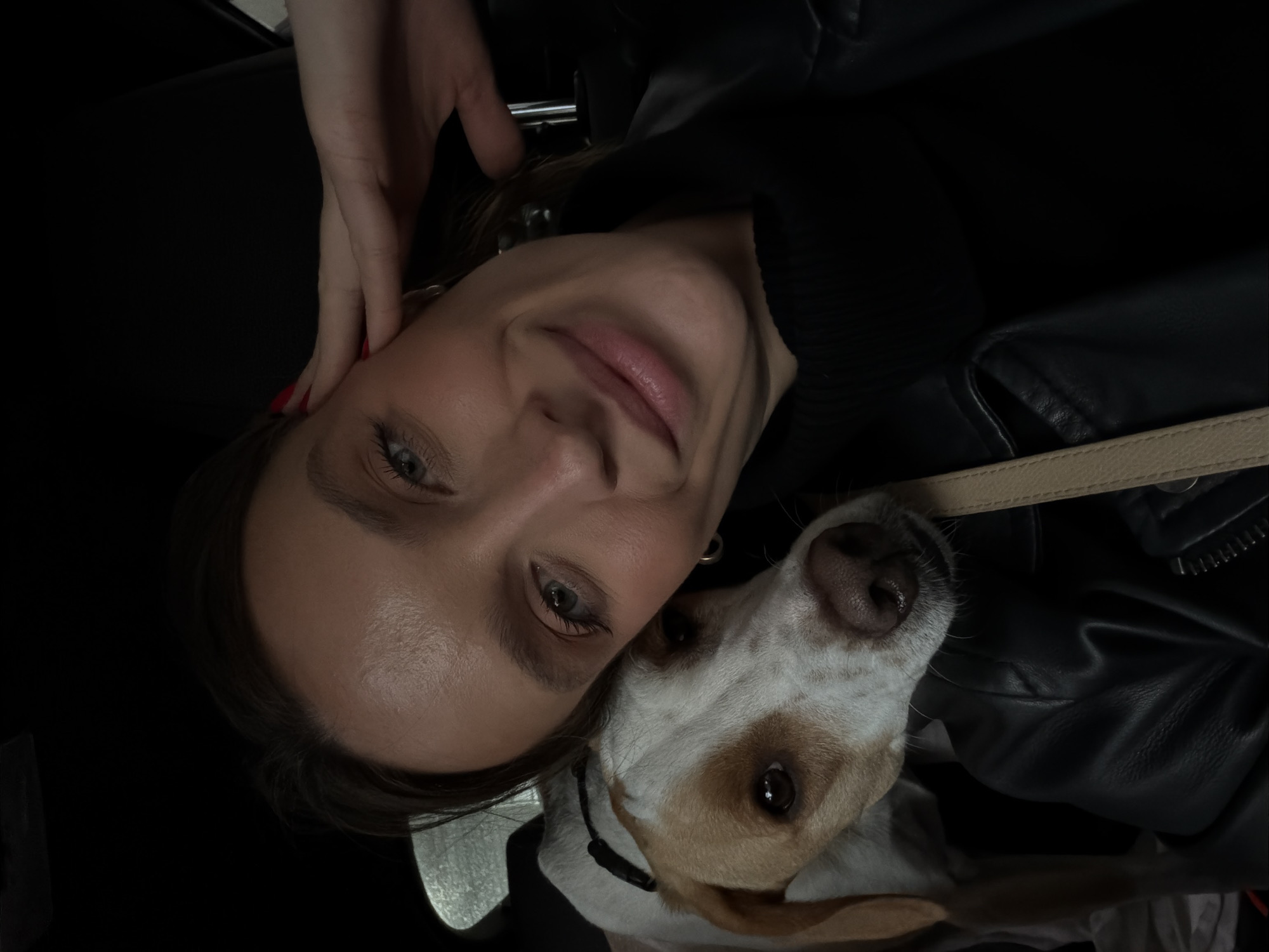 A close-up selfie captures an adult female and a domestic dog inside a vehicle in Budva, Montenegro. The female, appearing Caucasian with dark hair and blue-green eyes, occupies the right side of the frame, wearing a black turtleneck and a black leather jacket. Her right hand, exhibiting red nail polish, is positioned with fingertips touching her right temple. A subtle smile is present on her lips, and a gold hoop earring is visible in her left ear. A beige strap is visible across her right shoulder. A white and light brown bi-colored dog, with brown eyes and a dark brown nose, rests its head against the woman's left shoulder and chest area, facing the camera. The dog is equipped with a dark-colored collar. The interior environment of the vehicle is dimly lit, with a reflective surface, possibly a car window, visible in the upper left quadrant.