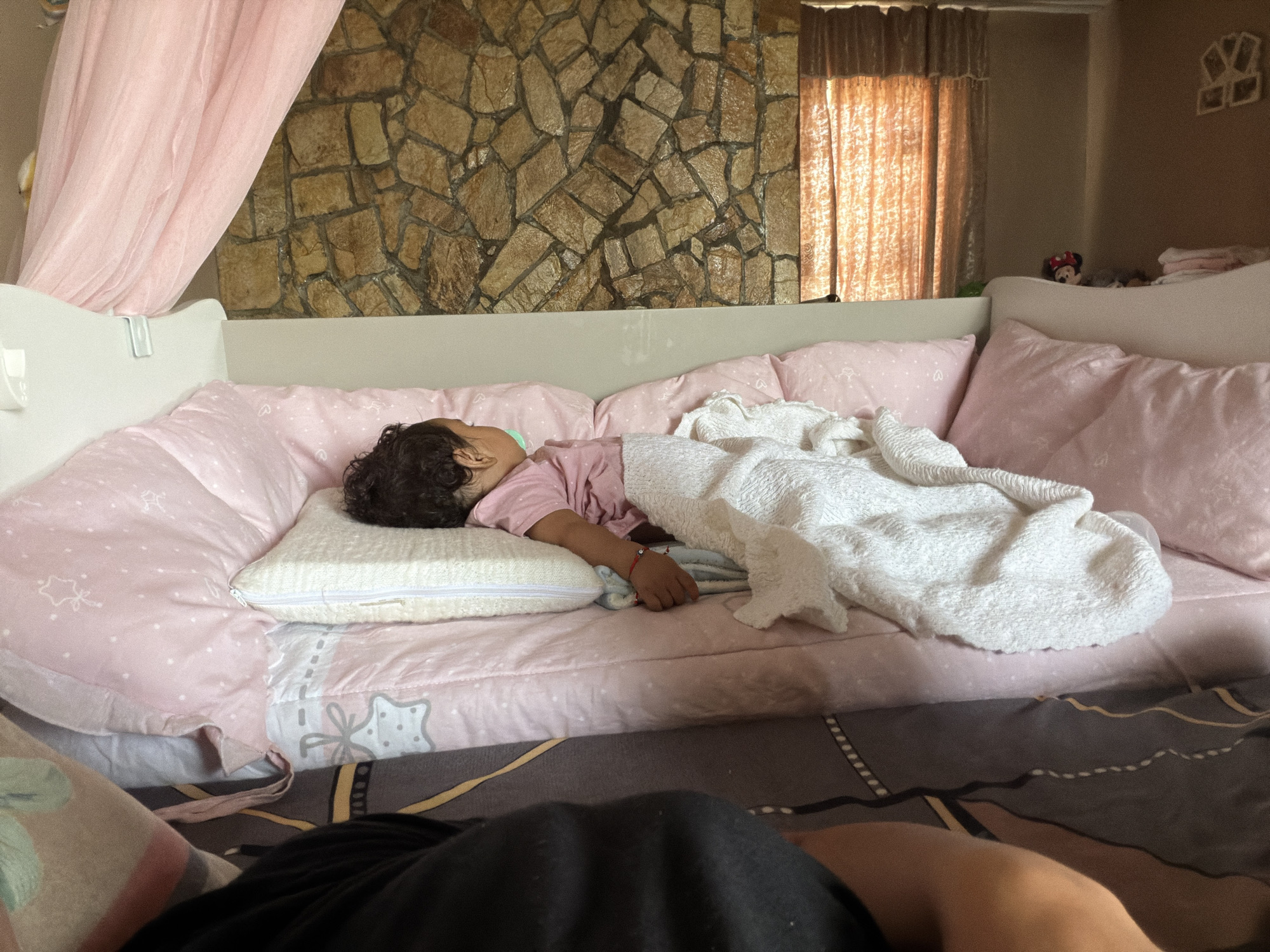 In a bedroom in Razhevo Konare, Bulgaria, a young child is sleeping soundly on a pink-covered bed. The child, wearing a pink shirt, is nestled on a small white pillow and covered with a white textured blanket. The bed is adorned with pink pillows featuring white star patterns. A pink sheer canopy drapes from above the bed. Behind the bed, a wall is covered with rough-hewn, irregular tan stones, and to the right, orange curtains cover a window, allowing soft light to filter into the room. In the background, a stuffed Mickey Mouse toy peeks over the edge of the bed. The foreground shows a person's legs and torso, indicating the photographer's perspective.