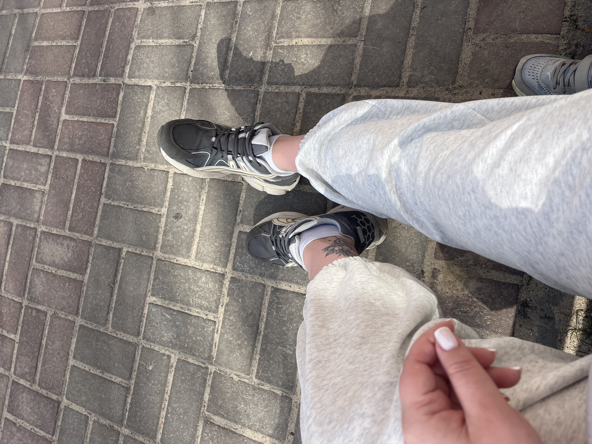 A first-person perspective photograph shows a person wearing light gray sweatpants and gray athletic shoes standing on a paved surface of rectangular, gray bricks. The person's left foot is visible with a tattoo on the ankle and a white sock peeking out of the shoe. The person's hand, with a light pink manicure, is also visible on the right side. Another foot wearing a different gray athletic shoe is partially visible in the upper right corner. The photo was taken during the day, with a shadow visible near the top of the image. It is not possible to verify the location is Usman, Russia based on the image alone.