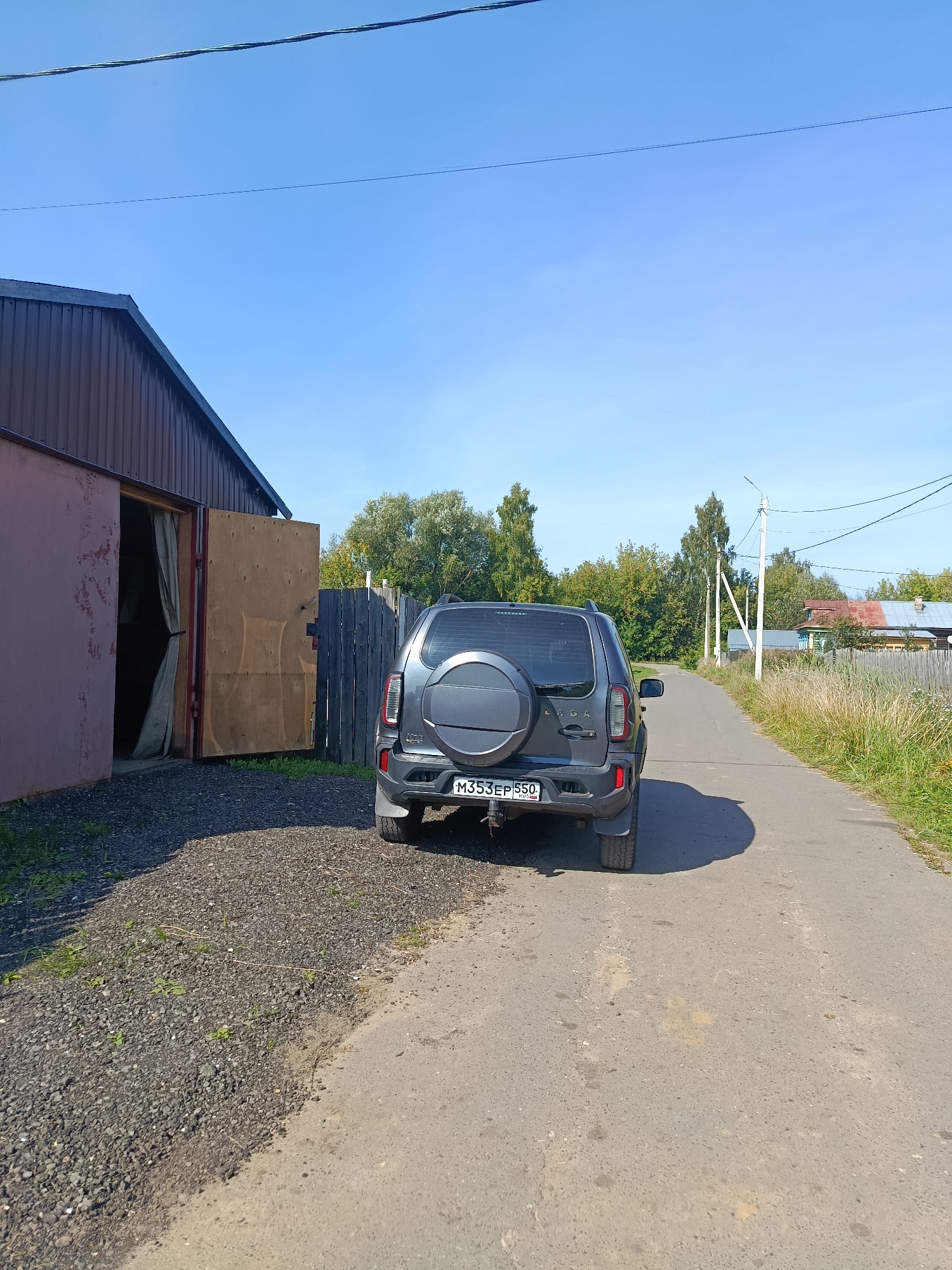 In a rural setting, a dark grey Lada Niva SUV is parked on a paved road, facing away from the viewer. The vehicle has a spare tire mounted on the rear hatch. To the left of the road is a gravel patch and an open-sided building with a dark brown corrugated metal roof and reddish-pink walls. The building's interior is partially obscured by a grey curtain. A wooden gate is partially visible to the right of the building. The road continues into the distance, lined with grass and trees. In the background, several houses with colorful roofs are visible, along with utility poles and power lines. The sky is clear and blue.