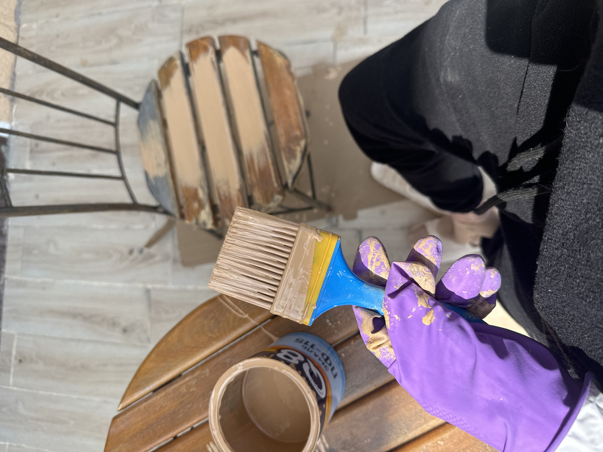Here's a detailed description of the image:
The image shows a first-person view of someone painting outdoor furniture. The location is in the city of Береговой сельский совет, Ukraine.
The person is wearing purple rubber gloves with paint splatters and is holding a paintbrush with a blue handle, yellow insert, and tan bristles. The bristles are coated in tan paint. The paintbrush is positioned above an open can of tan paint. The paint can has Cyrillic text on the label.
Below the paint can is a small, round, wooden table. The table is made of wooden slats that are painted tan. The table is sitting on a tiled surface with a wood-grain pattern.
Behind the table, a chair with a metal frame and wooden slats on the seat is partially painted tan.
To the right, the person is wearing black clothing and white shoes. The entire scene is lit by natural sunlight.