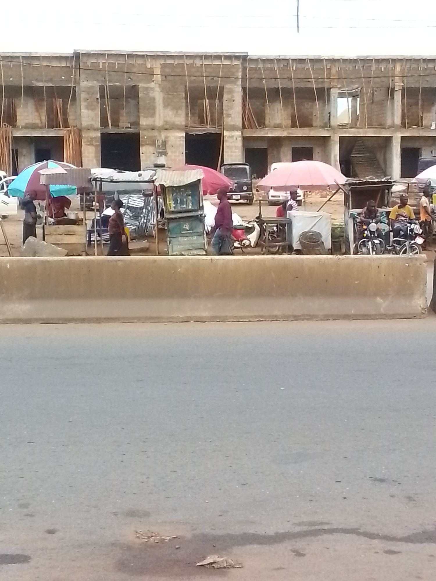 The image depicts a street scene in Ihiala, Nigeria. A wide concrete barrier divides the foreground road from the midground activity. Behind the barrier, vendors have set up stalls under colorful umbrellas, selling goods. Several people are visible, including vendors and customers. There are also motorcycles parked near the stalls. In the background, a partially constructed two-story building is visible. Electrical wires run across the top of the frame.