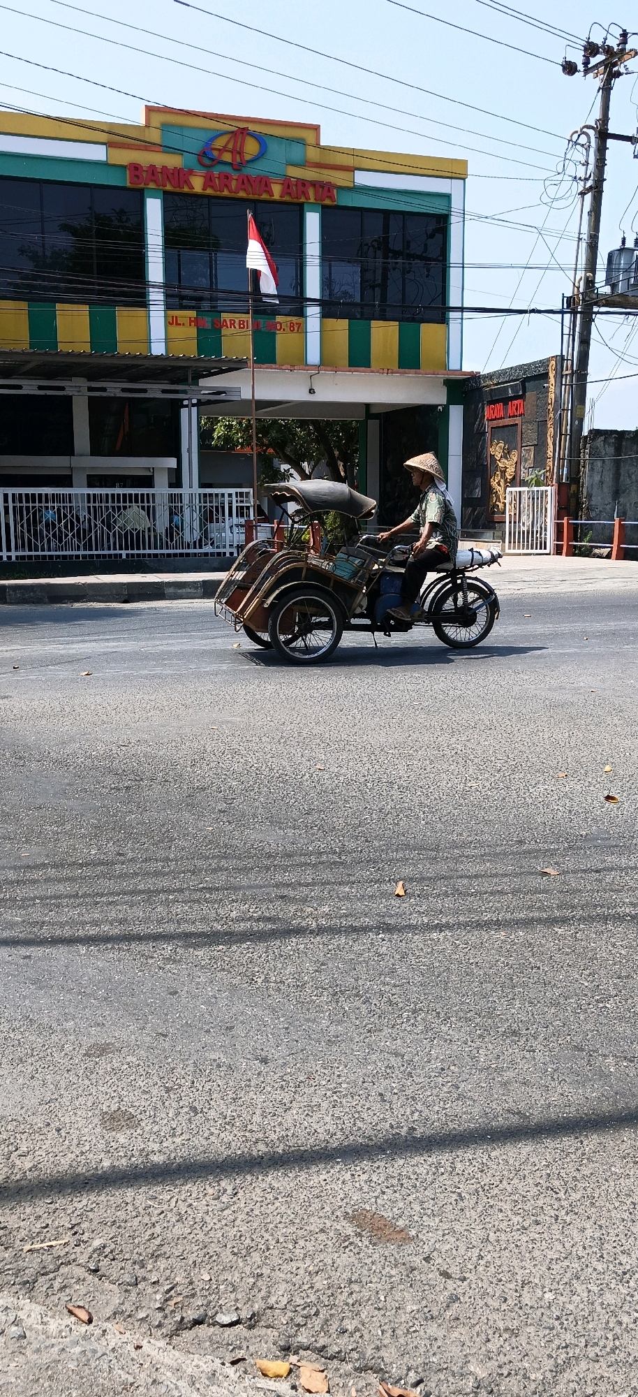 A pedicab driver, wearing a conical hat and a patterned shirt, operates a three-wheeled vehicle on a paved road in Kebumen, Indonesia. The pedicab, with a covered passenger area, is moving from left to right. In the background, a building with colorful stripes in green, yellow, and white displays the text "BANK ARAYA ARTA." An Indonesian flag is prominently displayed on a flagpole in front of the bank. The scene is illuminated by bright sunlight, casting shadows across the road. The environment appears to be an urban street, with power lines visible overhead and a concrete barrier along the roadside.
