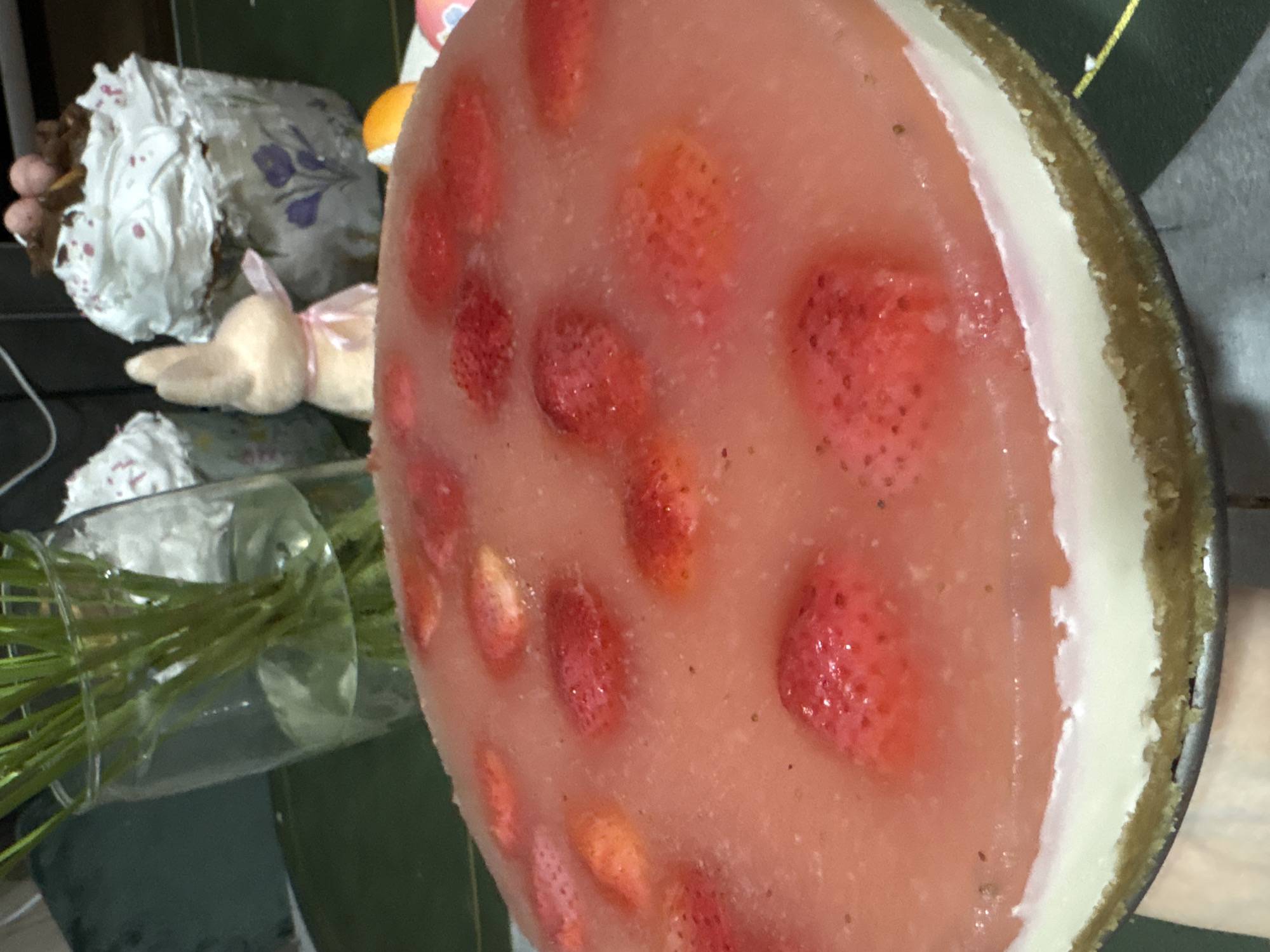 A close-up, slightly angled shot depicts a strawberry cheesecake with a gelatin topping and a graham cracker crust. Fresh strawberries are embedded in the gelatin. The cheesecake sits on a surface with other objects in the background, including a vase with green stems, a stuffed bunny, and floral-patterned fabric. The location is unidentified, and there is no explicit connection to Chaikovsky, Russia, in the image itself.