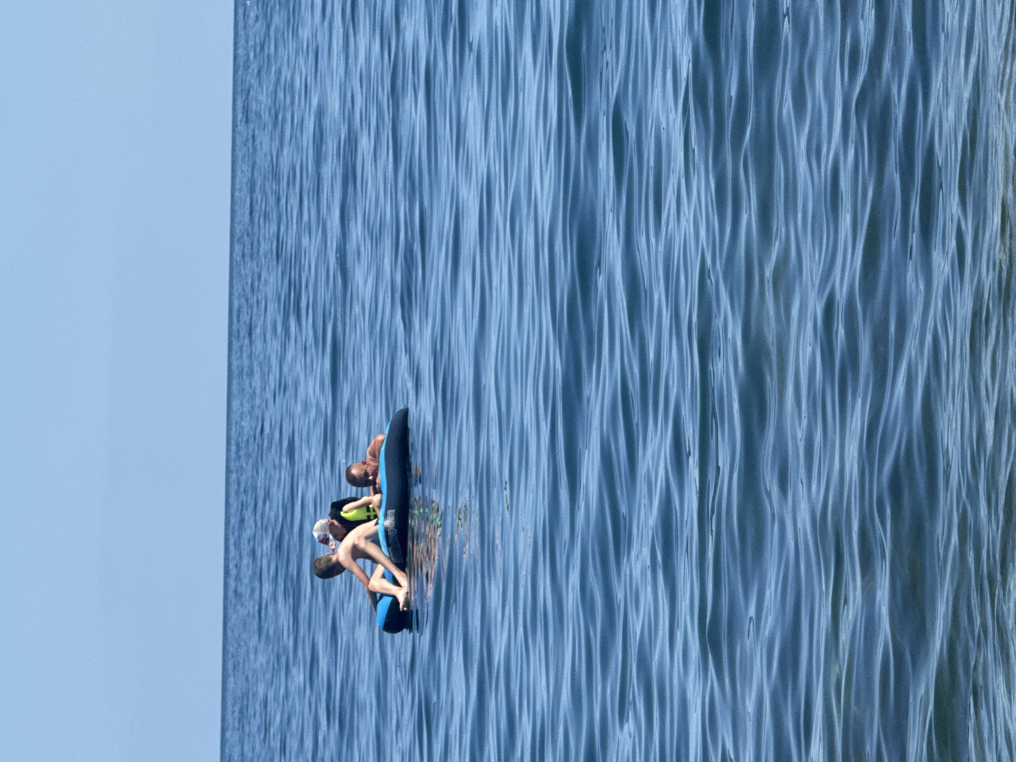 The image depicts three individuals, two children and one adult, on an inflatable raft in the open sea. The water is blue and moderately rippled. The sky is a clear, pale blue. One child, wearing a white baseball cap and a yellow life vest, is seated facing left, with his legs extended. Another child, bare-chested and wearing a white baseball cap, is seated behind the first child, also facing left. An adult male, bare-chested, is lying down on the raft, facing towards the right of the image. The raft is dark blue and black and appears to be floating on the surface of the water. The scene suggests a recreational activity in a marine environment. This specific location is not identifiable within the city of Utios, Ukraine, as there are no discernible landmarks or geographical features that would confirm the location.