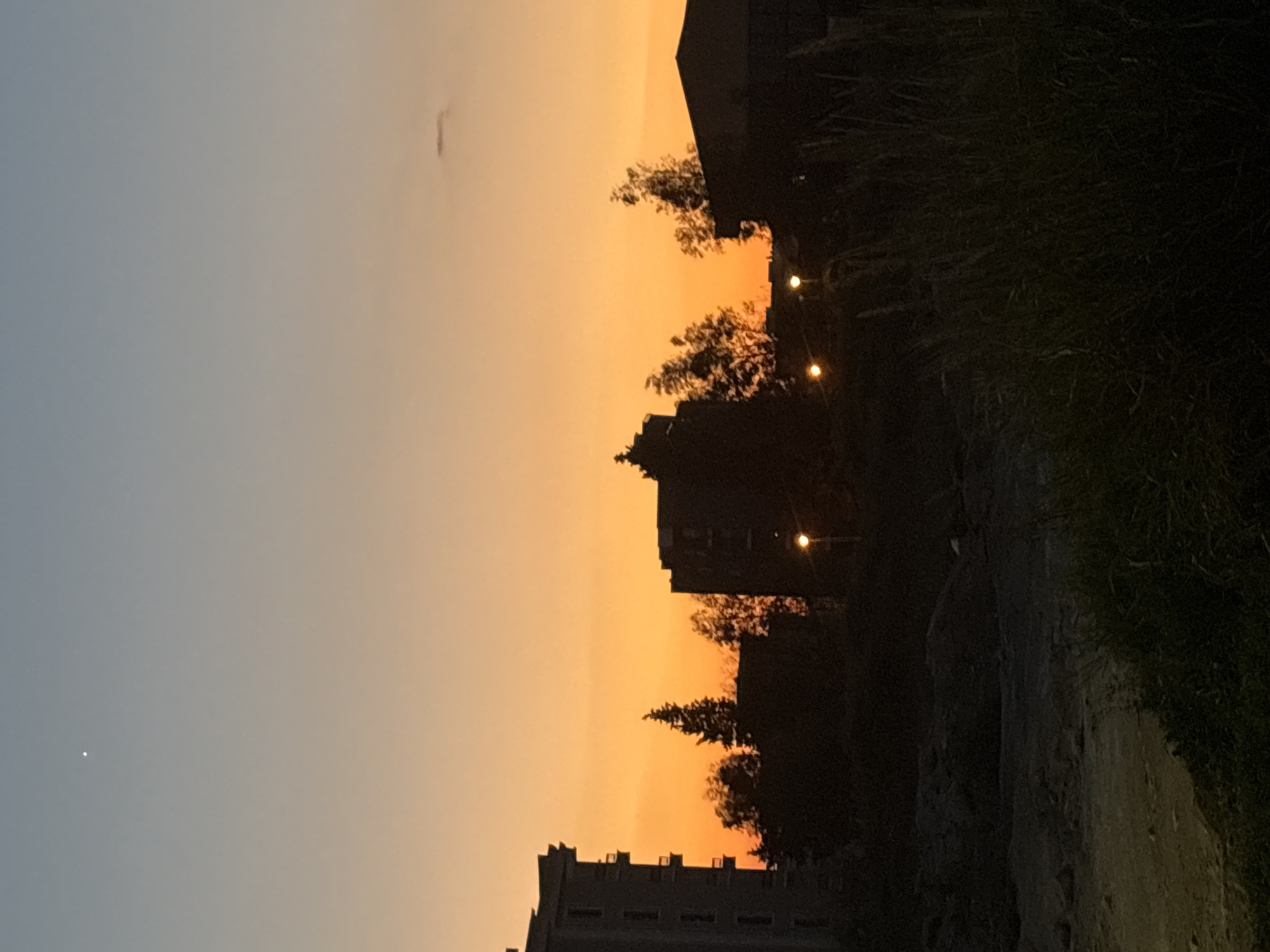 The image depicts a view at sunset in the city of Bar, Montenegro. The sky transitions from blue to orange. Silhouettes of buildings, trees, and vegetation are visible against the bright sky. There are several bright lights (possibly streetlights) interspersed among the buildings and trees. A path is present in the foreground.