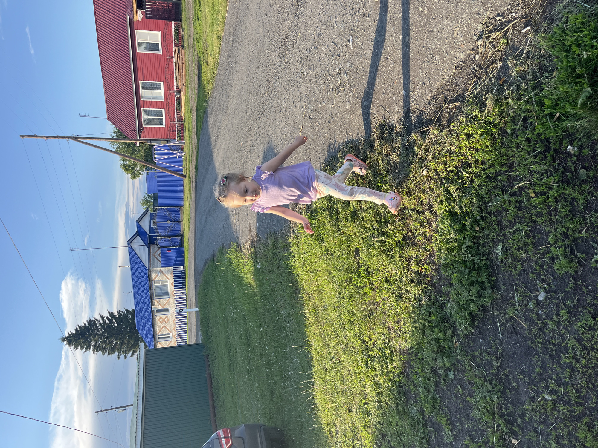 In Mal'tsevo, Russia, on a sunny day, a young blonde girl with pigtails adorned with watermelon-shaped clips runs on a gravel road. She is wearing a purple t-shirt with ruffled sleeves and patterned leggings, along with pink clogs. The girl is in motion, with her arms outstretched and her right leg forward as if mid-stride. To her left, a patch of green grass is illuminated by the sun, contrasting with the darker earth. Behind her, a road stretches towards the background, where a red-roofed house with white trim and windows stands prominently. Further left, a green building is visible, and beyond that, a house with a blue roof and white fence. A tall evergreen tree is situated near the blue-roofed house, and utility poles with wires run across the bright blue sky dotted with fluffy white clouds. The overall scene depicts a rural residential area on a clear day.