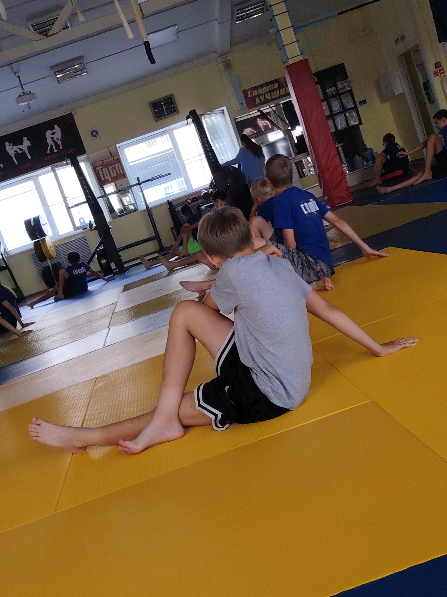 The image depicts a group of young individuals, likely boys, engaged in what appears to be a martial arts or athletic training session in a gymnasium. They are positioned on a yellow and blue mat, common in dojos or sports halls. Several individuals are seen sitting or stretching, with one boy in the foreground wearing a grey t-shirt and black shorts, his legs extended. Another boy in a blue t-shirt is visible in the background. The environment includes athletic equipment such as weight plates and a rowing machine, as well as gym banners with silhouettes of combatants and text in Russian, suggesting a sports club or school. The gym has a clock on the wall, fluorescent lighting, and windows with blinds. A sign above a doorway reads "СПАРТА ЛУЧШИЙ" which translates to "SPARTA THE BEST". This scene is likely located at the "Sparta" sports complex in Chaikovsky, Russia, a city known for its sports facilities, particularly for combat sports.